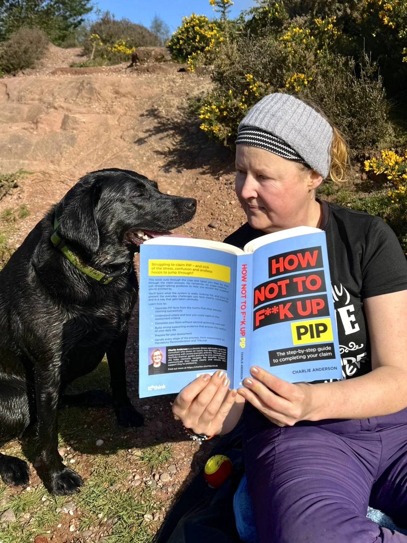 A woman with a gray headband sits outdoors reading a book titled 'How Not to F**k Up PIP' while a black dog with a green collar looks at her. The background features shrubs, yellow flowers, and a dirt path.