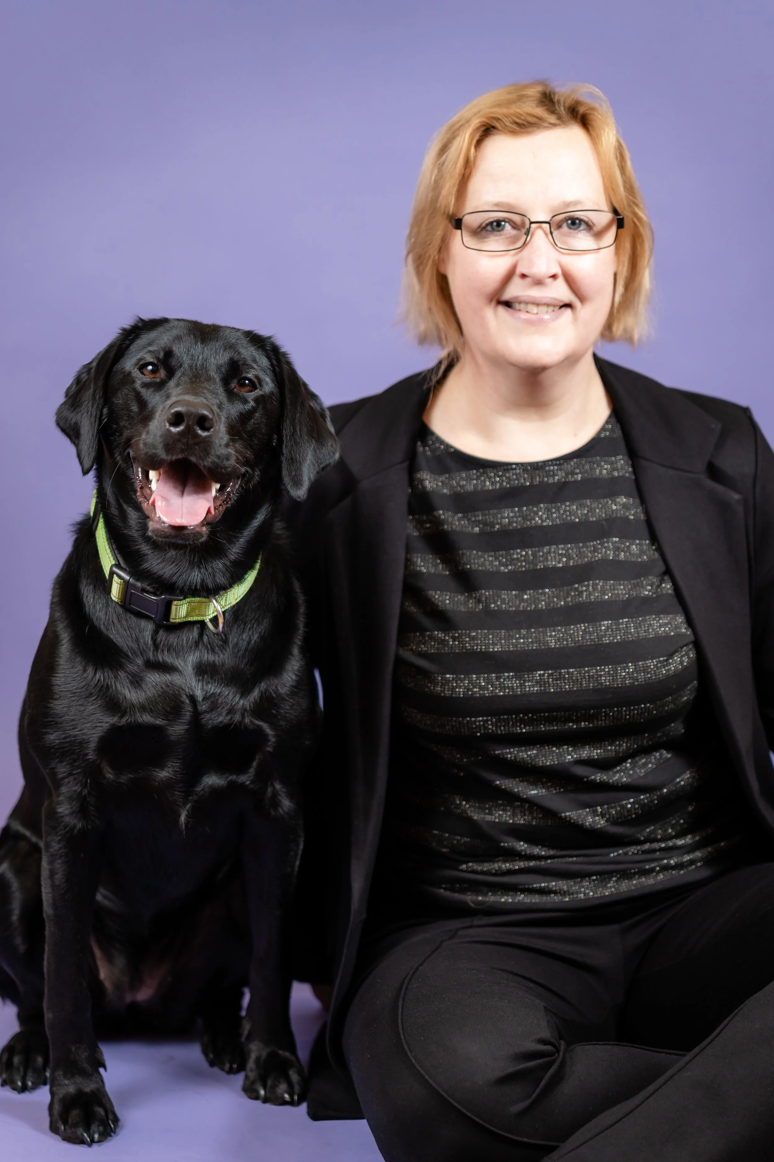 A woman wearing glasses smiling next to a black Labrador retriever with a green collar, both sitting against a purple background.