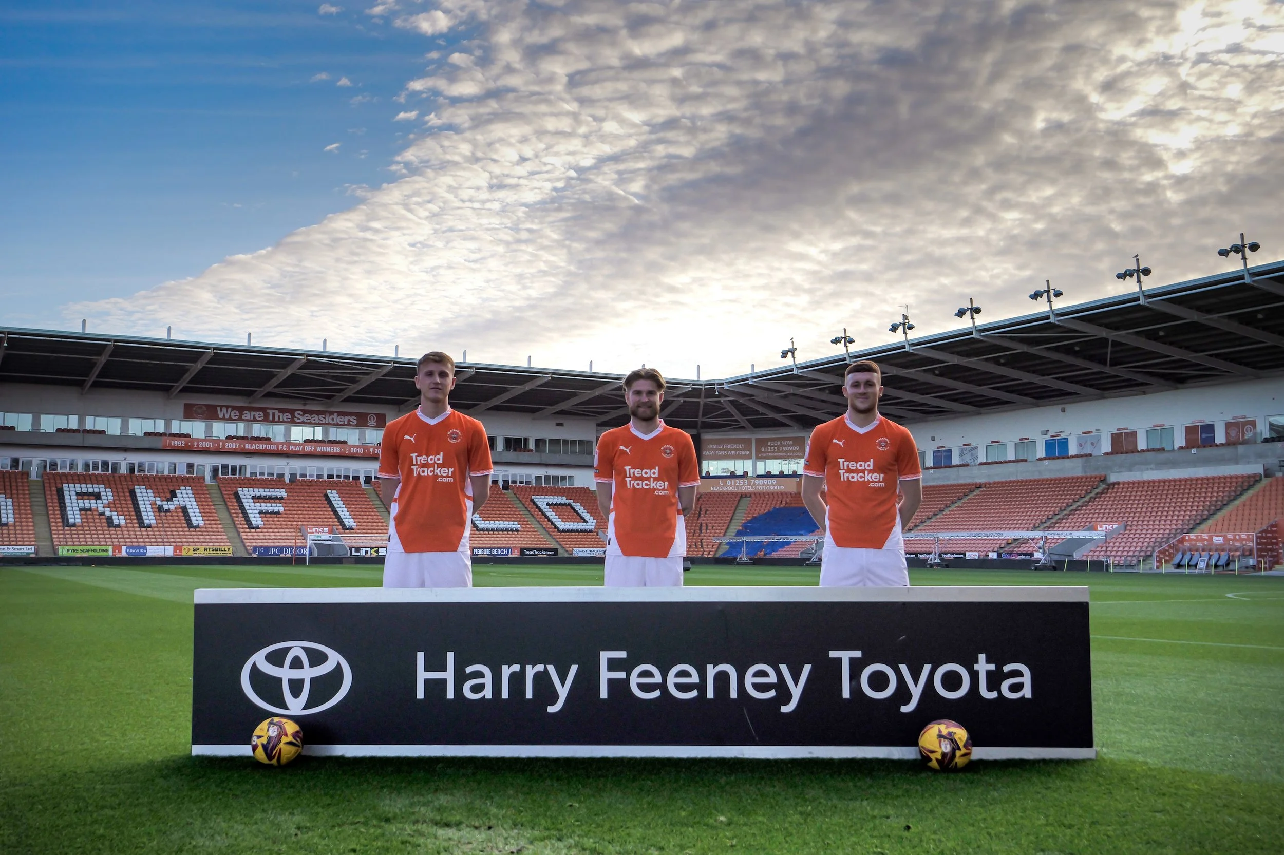 Three soccer players in orange jerseys standing on a soccer field, with stadium seating in the background and a blue sky with clouds overhead.