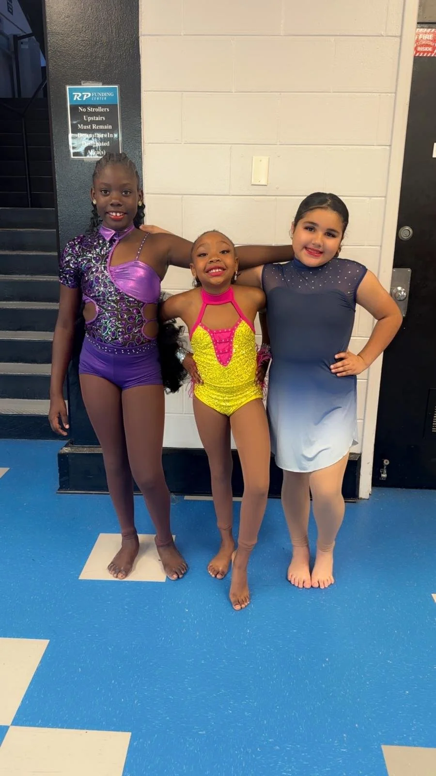 Three young girls dressed in colorful dance costumes standing together with their arms around each other in a hallway.