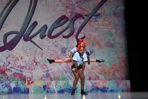 Two young girls performing a dance on stage, one girl supporting the other in a horizontal full split stunt.