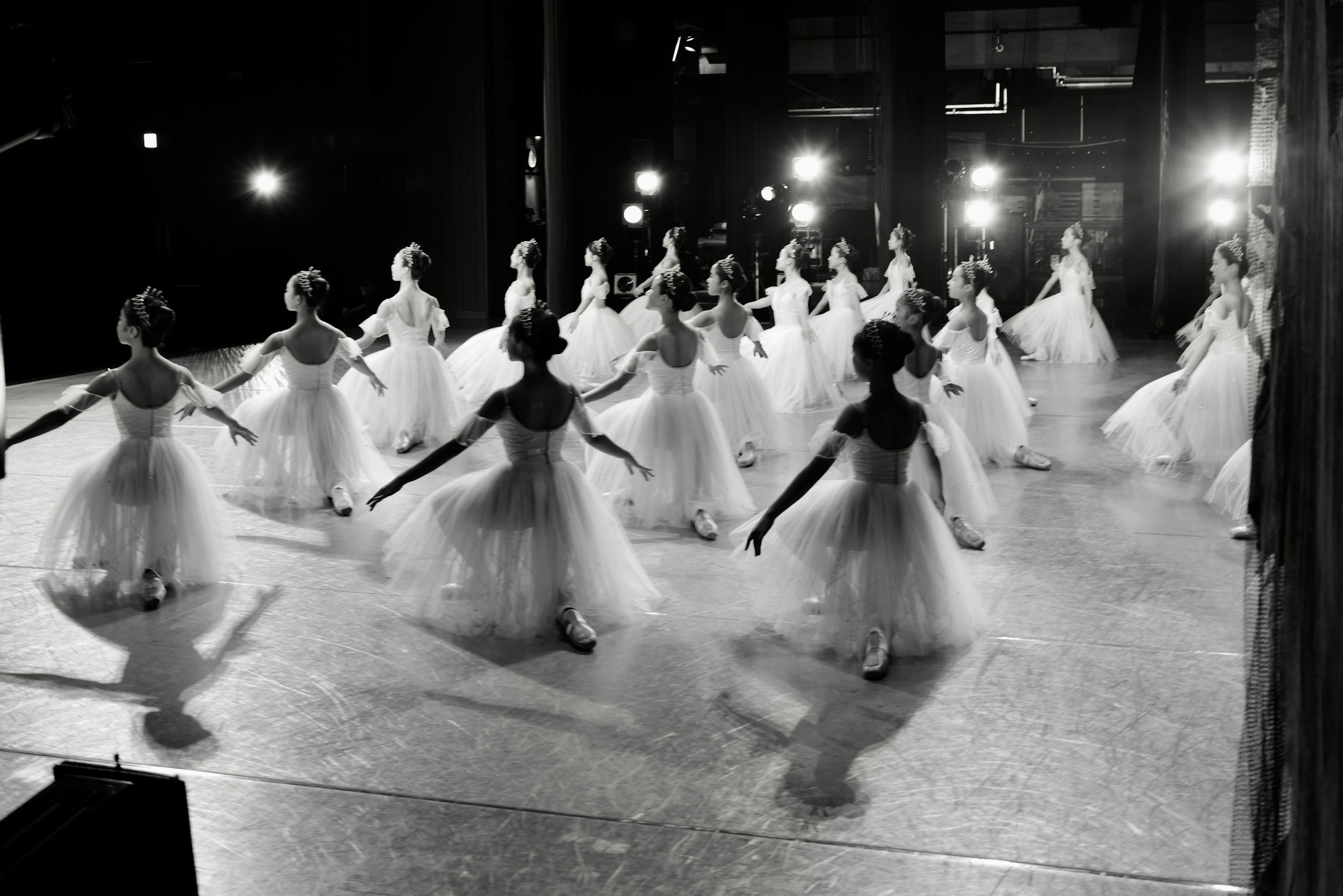 Ballet dancers in white tutus rehearsing on stage with bright lights and dark background.