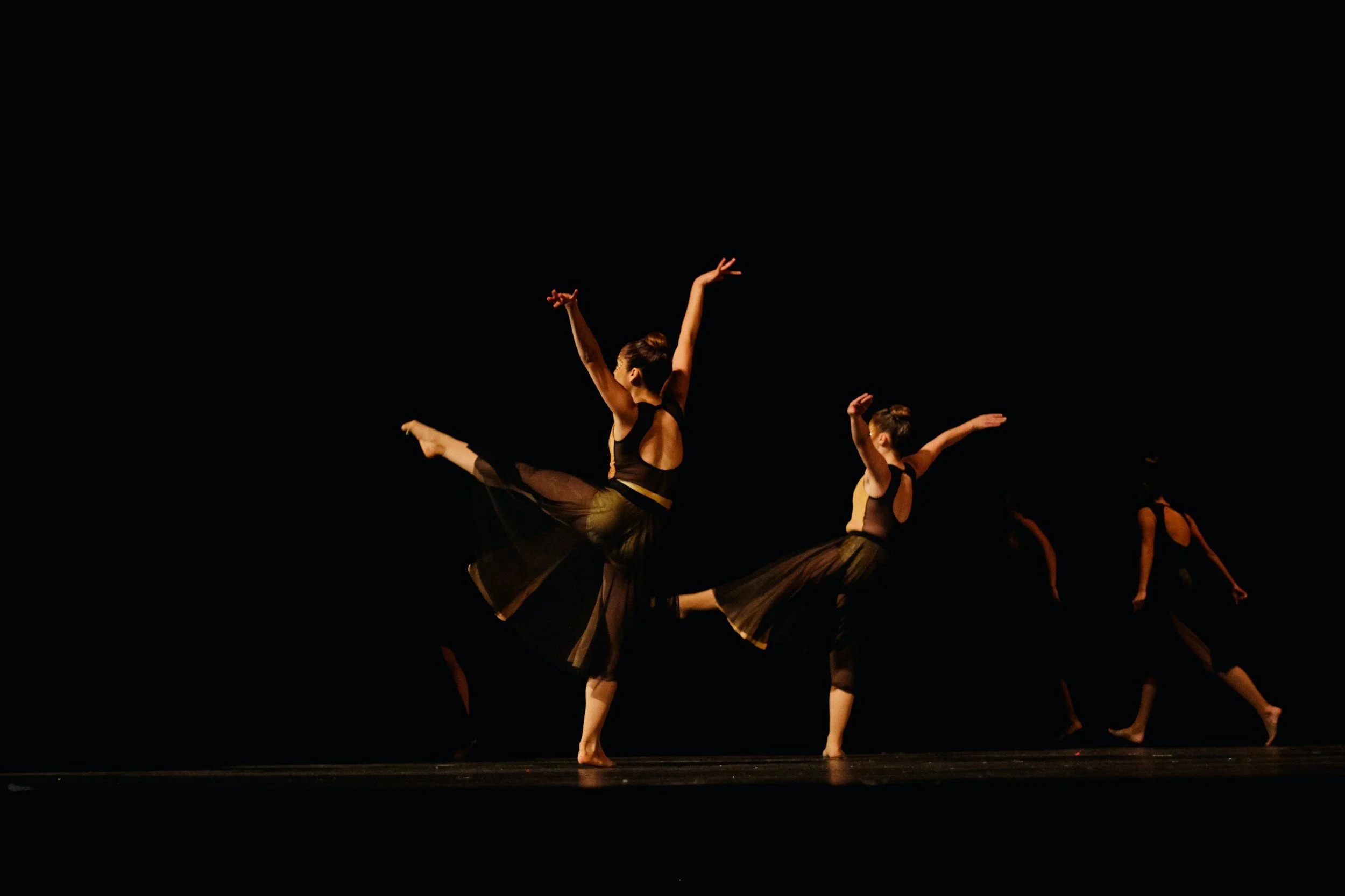 Group of ballet dancers performing on stage with black costumes, illuminated against a dark background.