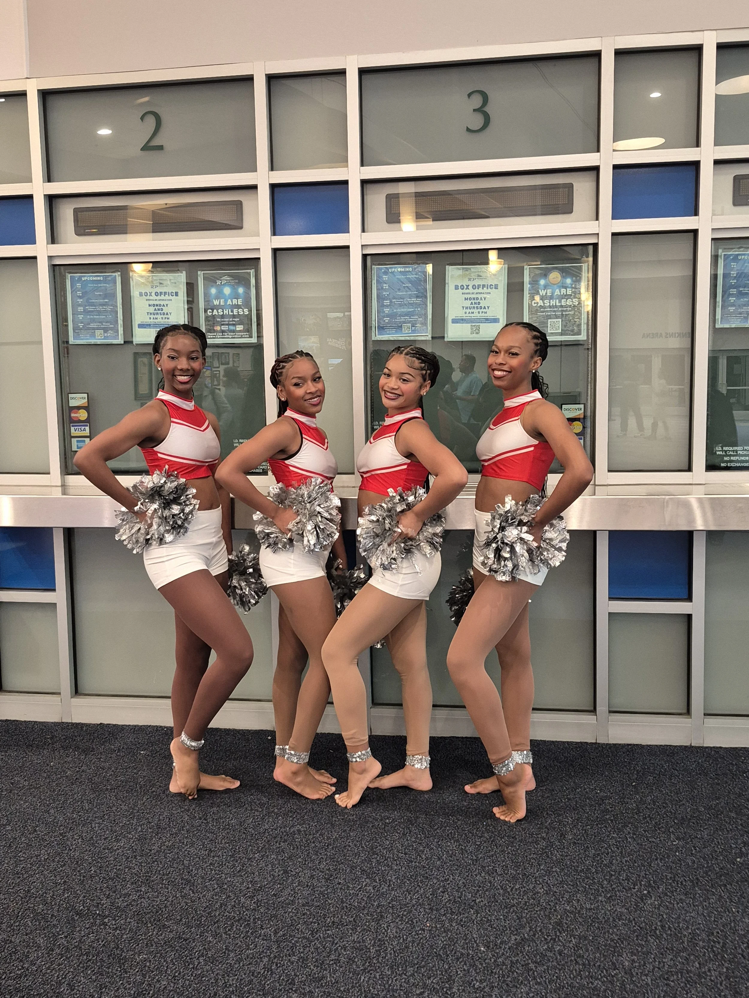 Four young women dressed in cheerleading uniforms pose in front of glass doors numbered 2 and 3, holding silver pom-poms, smiling, and standing on a dark carpeted floor.