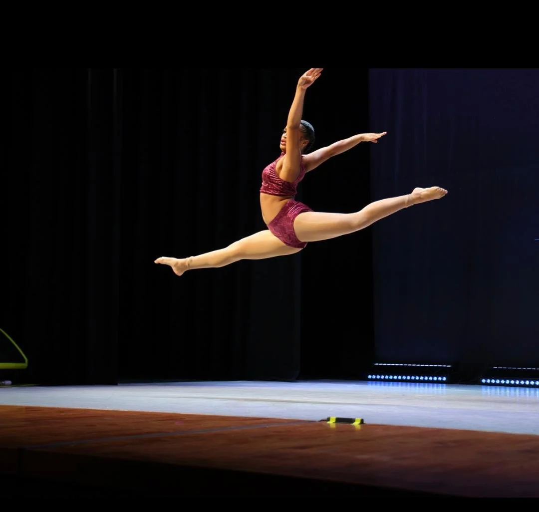 A female gymnast in a maroon athletic outfit performing a split leap in mid-air on a stage with a black background.