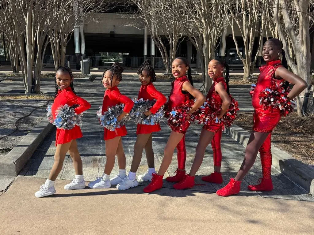 Six young girls dressed in red cheerleading outfits with pom-poms, standing in a line outdoors on a sidewalk with trees behind them.