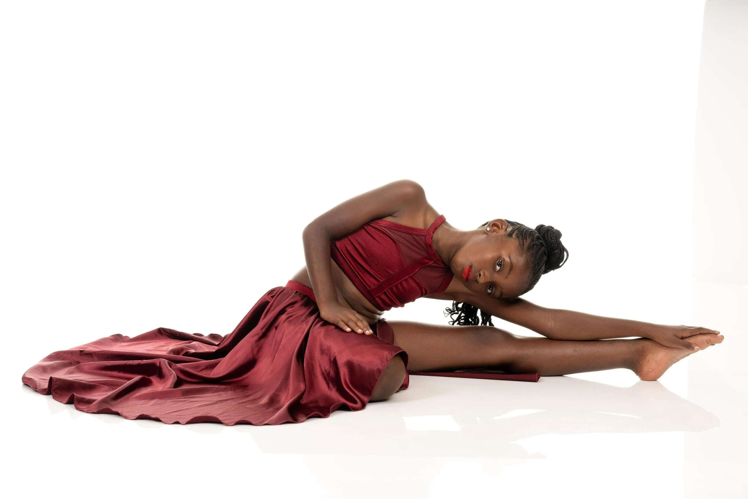 Our student/dancer Jade in a burgundy lyrical dress is performing a stretch on the floor, with her upper body turned to the side, head resting on her arm, and legs extended.