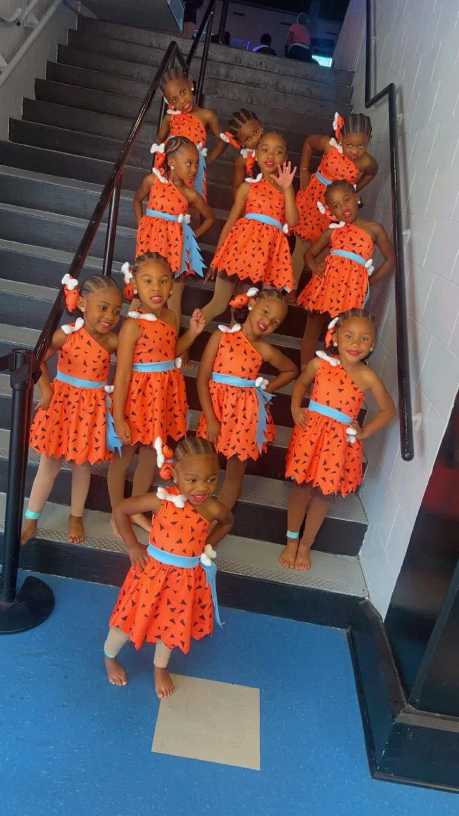 A group of young girls dressed in matching orange costumes with black triangle patterns, blue sashes, and white accessories, posing on a staircase indoors.