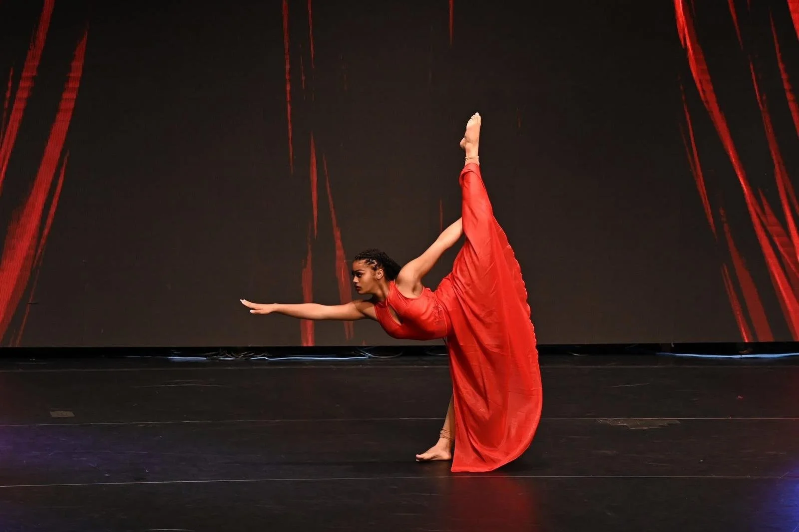 A female dancer in a red dress performing a standing split on stage with a black background and red stage curtains.