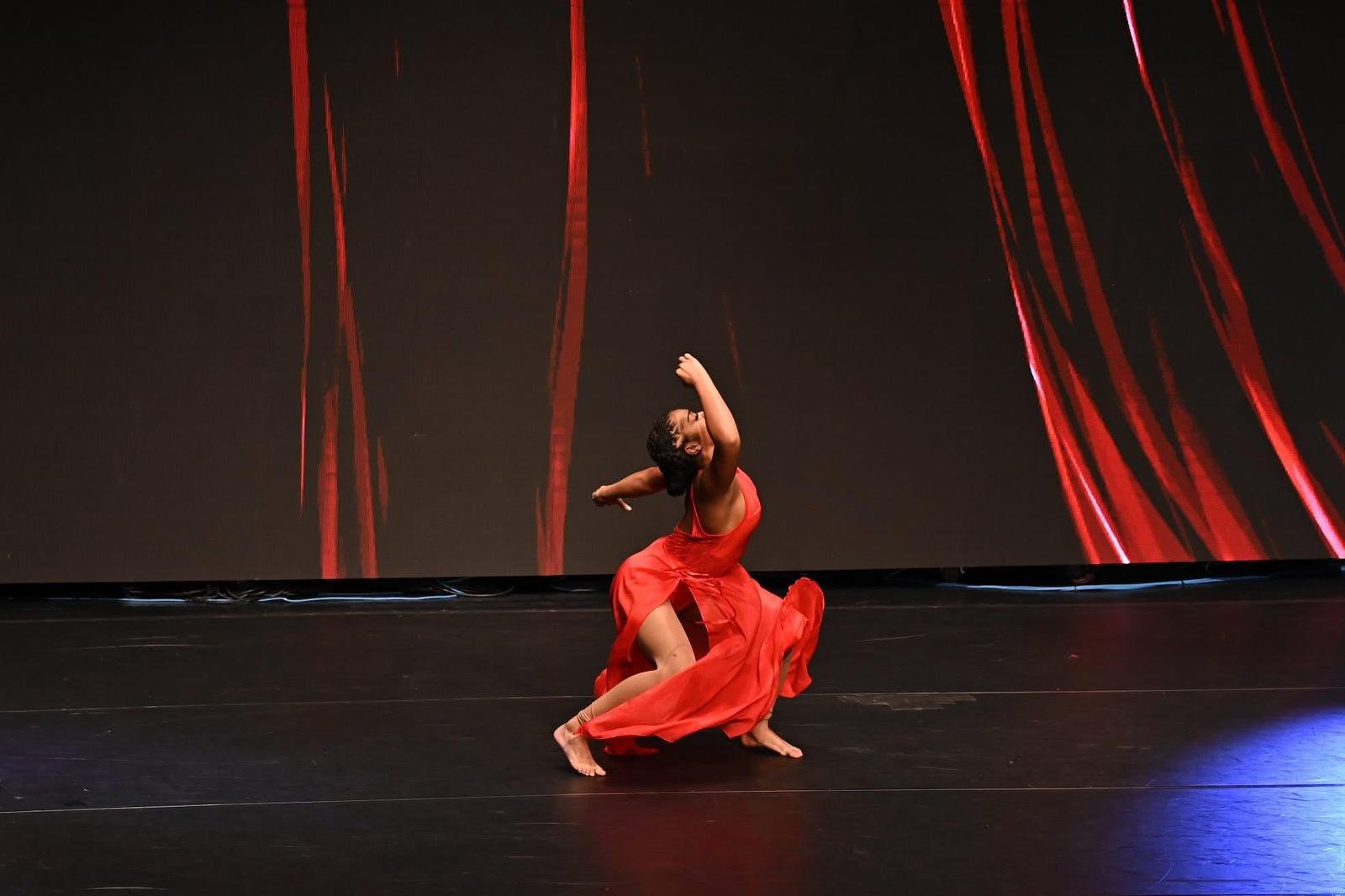 A female dancer in a flowing red dress performs on stage with a dark background and red streaks.