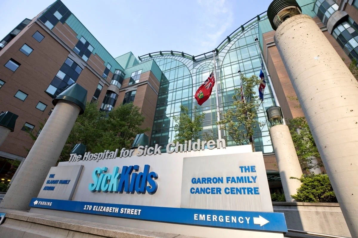 Exterior view of The Hospital for Sick Children, also known as Sick Kids, with a large sign displaying the hospital's name and related information. The building features a modern glass and brick design with flags flying above and trees in front.