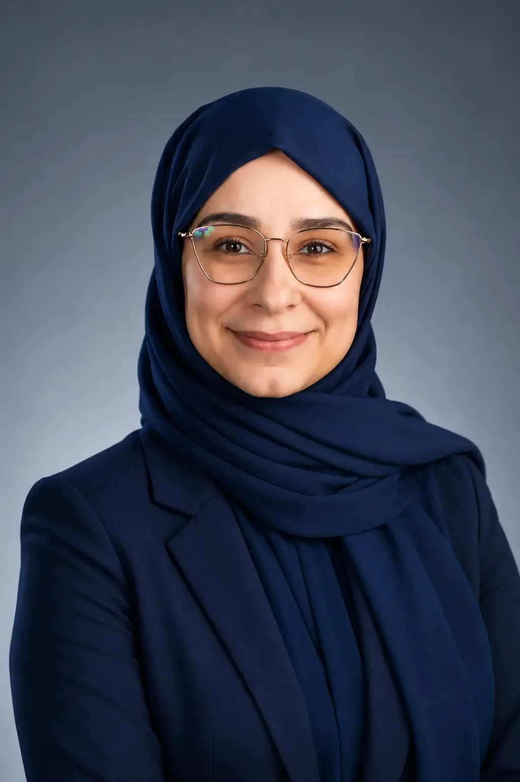 A woman wearing a navy blue hijab, glasses, and a matching blazer, smiling at the camera against a gray background.