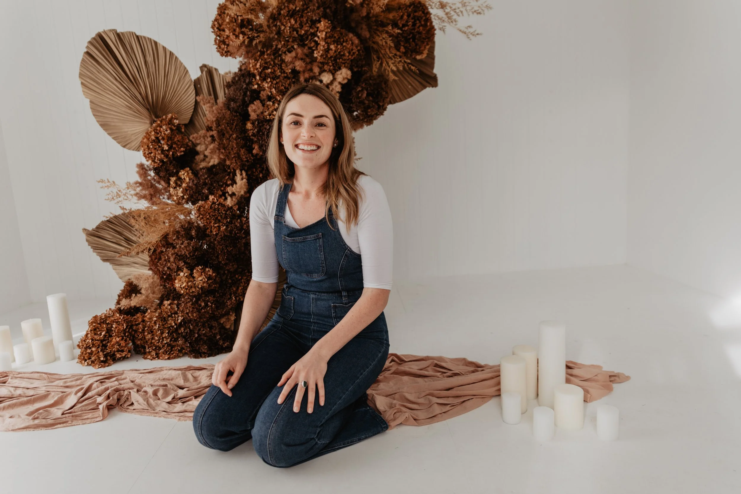 A woman with wavy brown hair smiling, kneeling on a peach-colored fabric, in front of a backdrop of dried flowers and large palm leaves, surrounded by white candles.