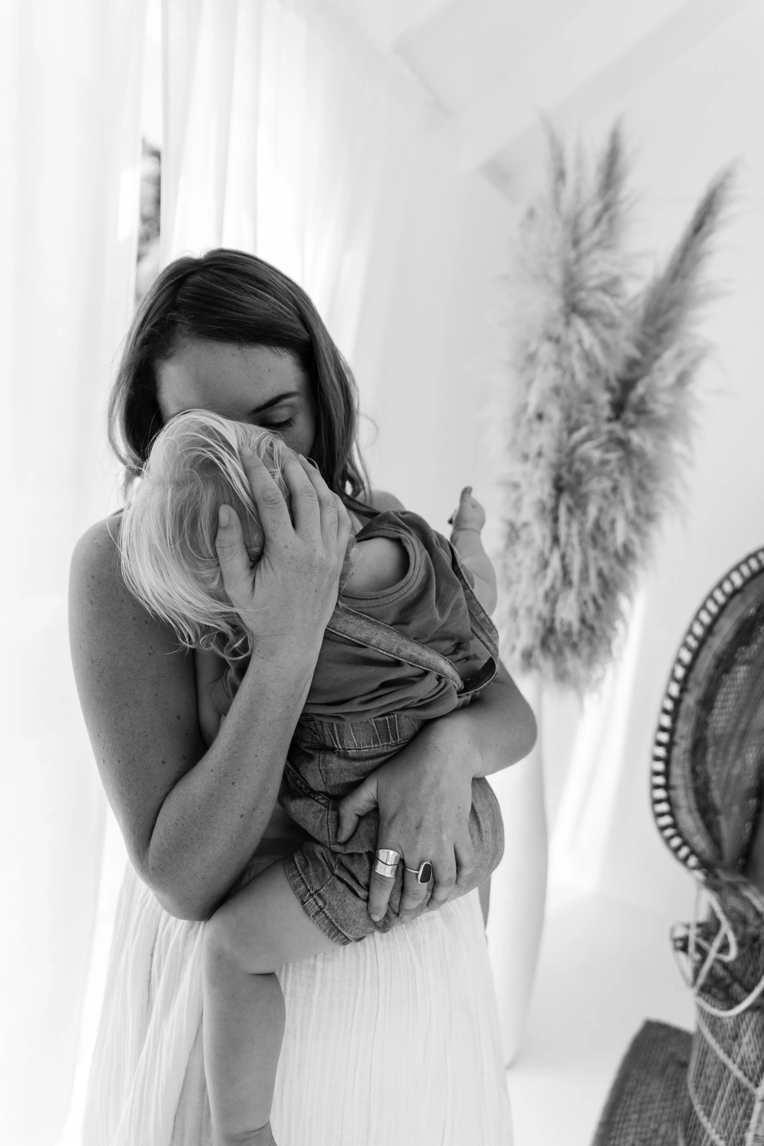 A woman holding a young child in a loving embrace indoors, with a white curtain, decorative flowers, and a basket with a blanket in the background.