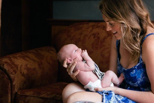 A woman holding a newborn baby on her lap, smiling and looking at the baby in a cozy indoor setting.