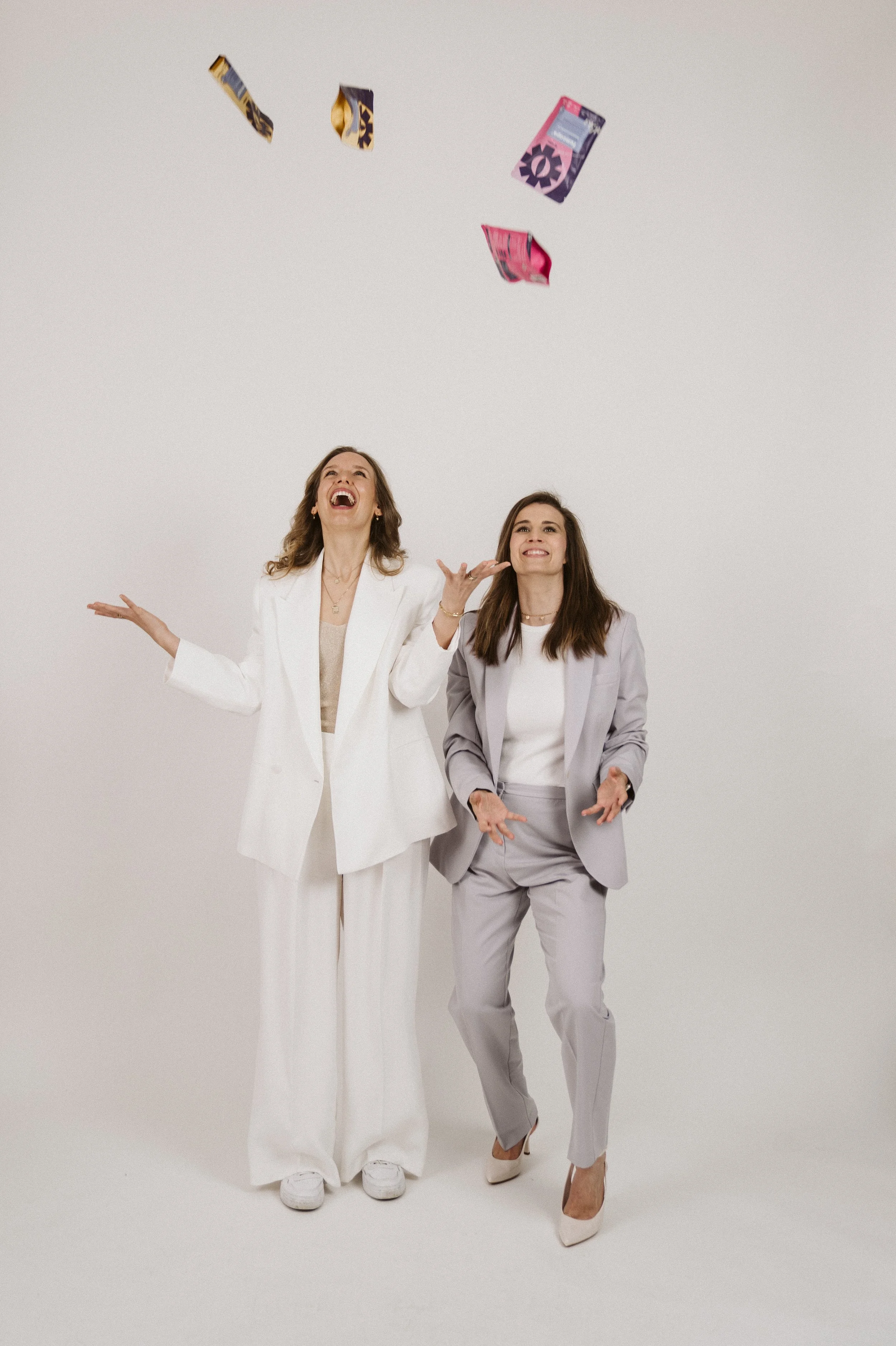 Two women standing against a plain light background, one laughing and throwing small snack bags in the air, both dressed in suits.