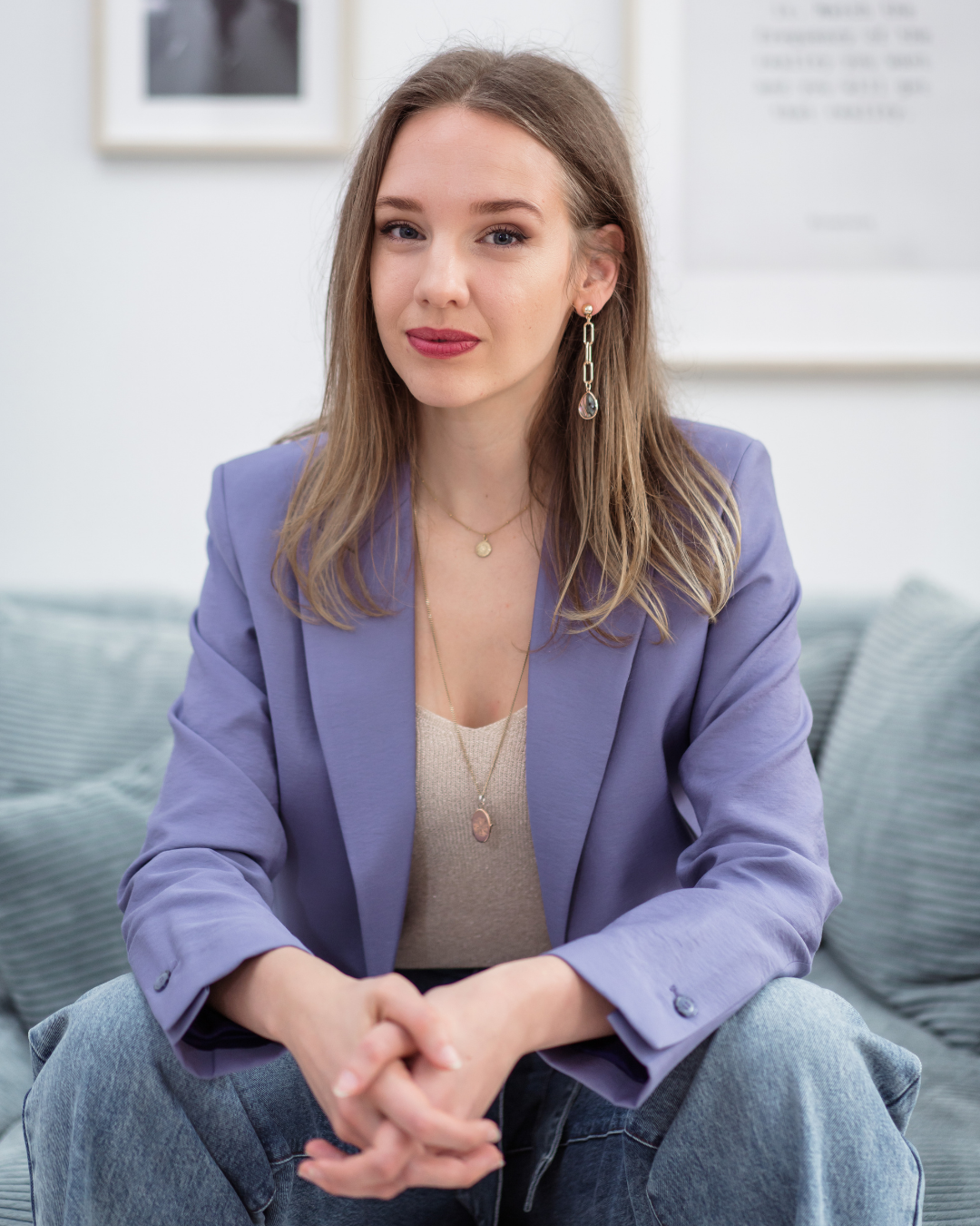 A young woman with light brown hair and blue eyes, wearing a lavender blazer, beige top, and jeans, sitting on a sofa with a neutral expression.