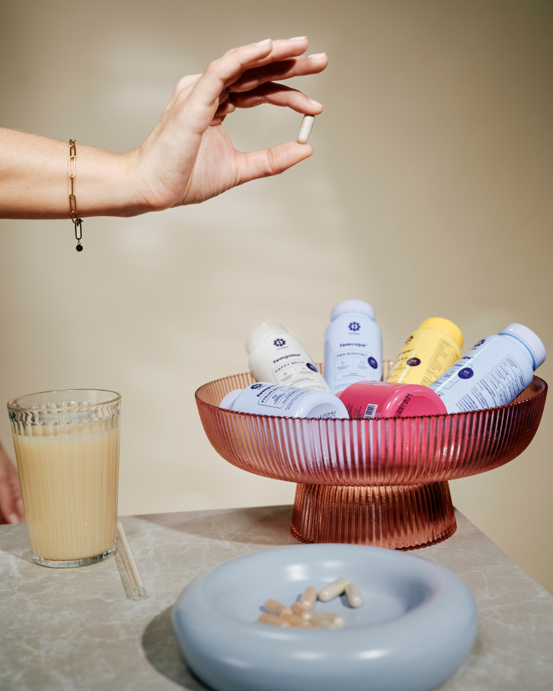 A hand holding a white capsule pill above a bowl of pills, with a glass of yellow liquid and a bowl of assorted medicines on a marble surface.
