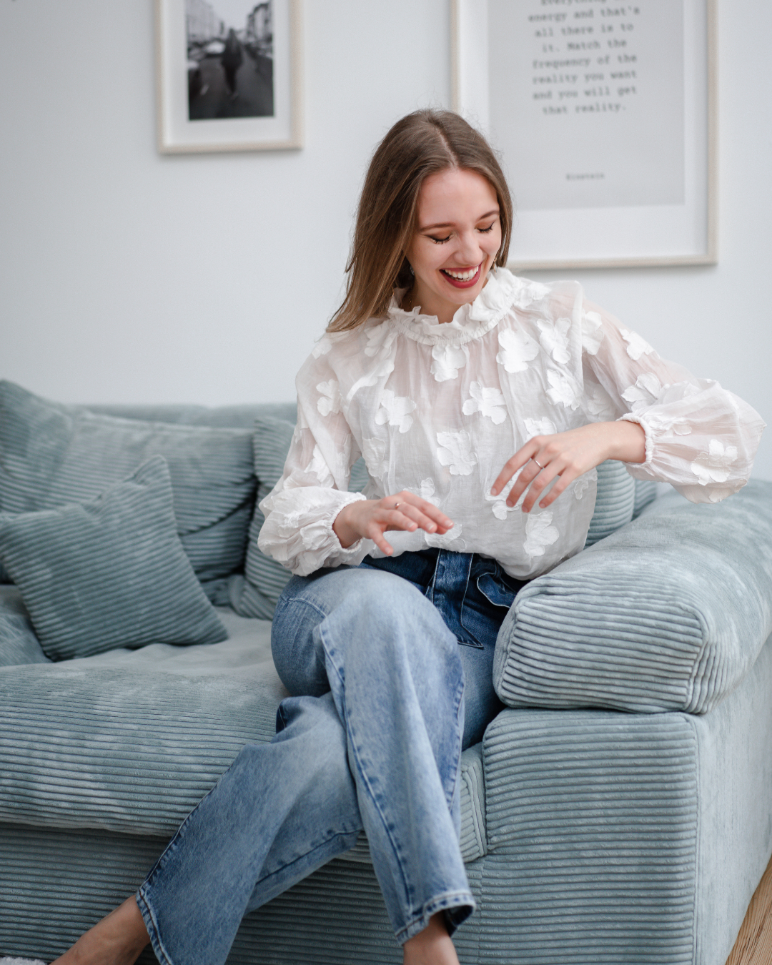 A young woman with long hair, wearing a white floral blouse and blue jeans, sitting on a light blue striped couch and smiling while looking at her wrist, in a cozy living room with framed pictures on the wall.