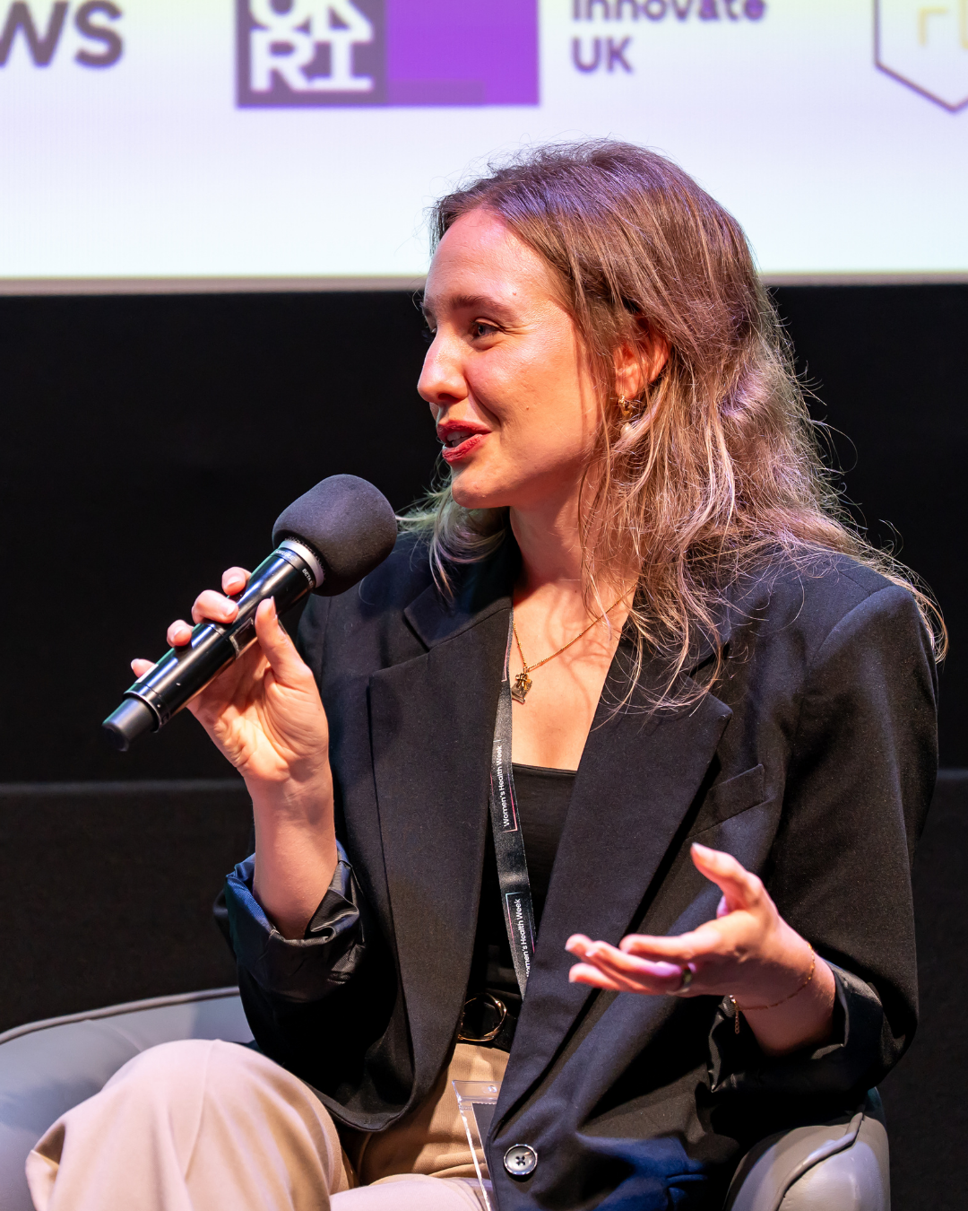 A woman with wavy brown hair speaking into a microphone during a panel discussion at an event. She is wearing a black blazer and beige pants.