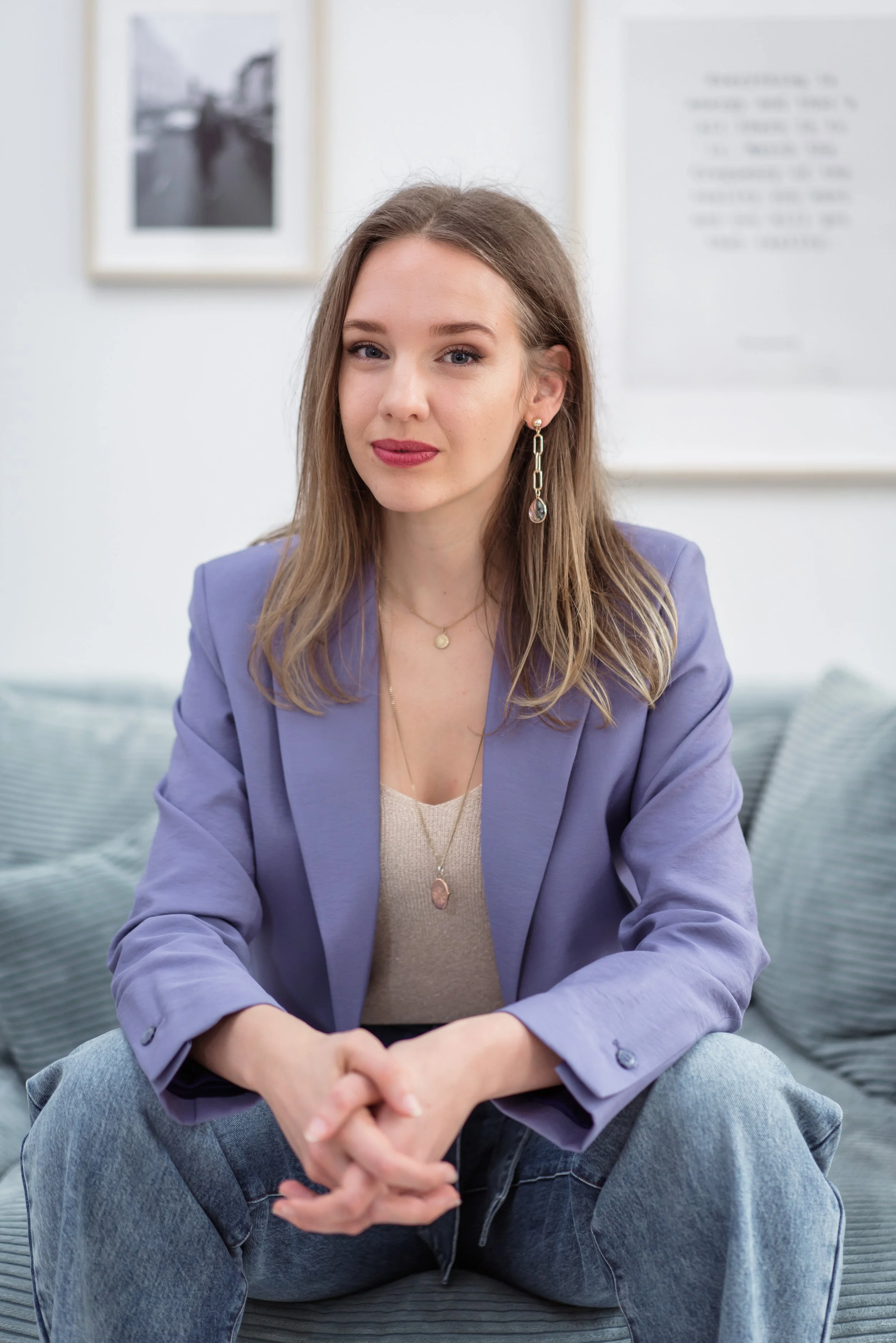 A young woman with light skin and long light brown hair sitting on a gray cushioned bench in a bright indoor space. She is wearing a lavender blazer, a beige top, gray jeans, and gold jewelry, including a chain necklace with a pendant and earrings. There are framed pictures and a quote on the wall behind her.
