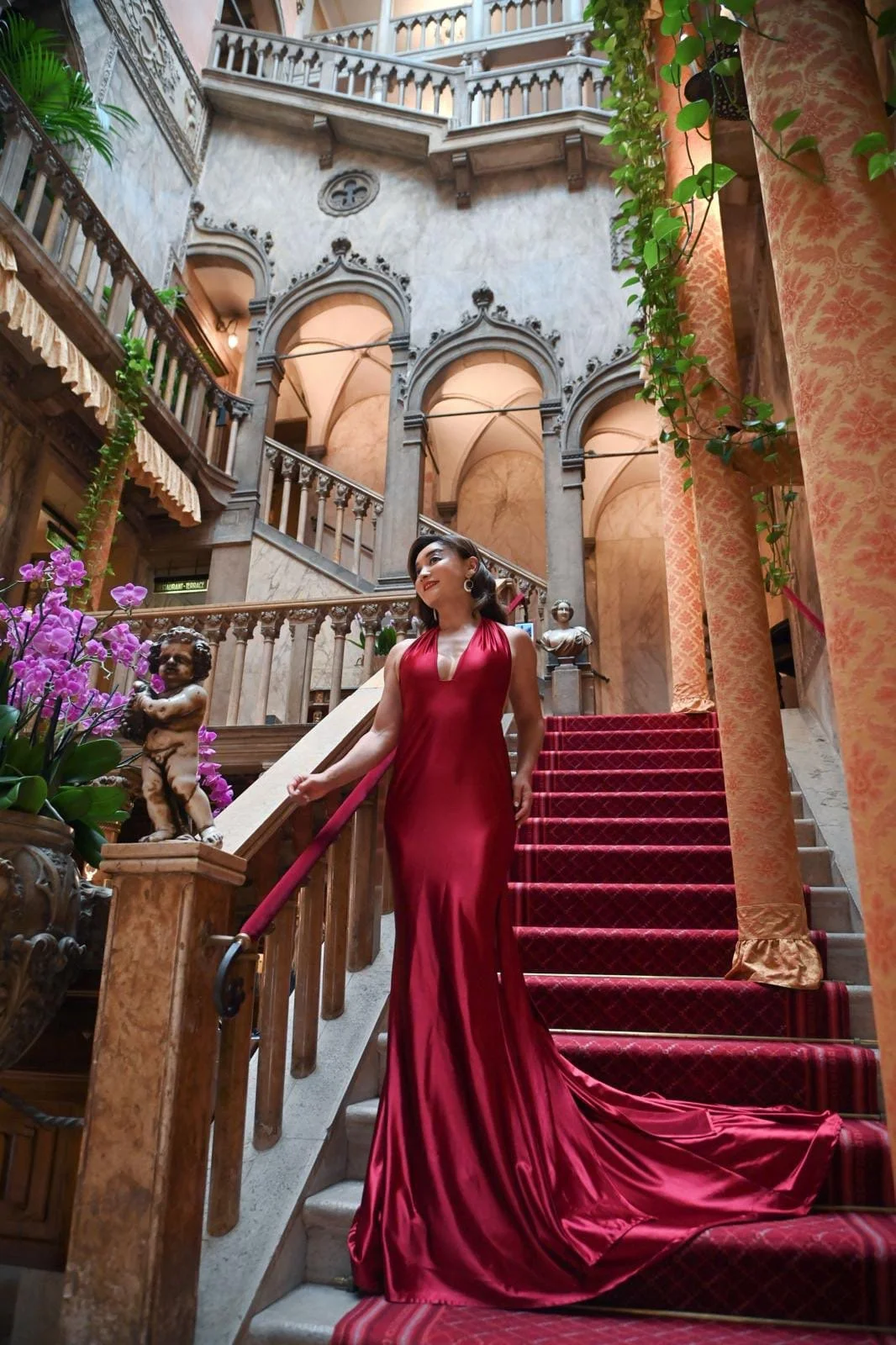 Winnie Wen in a red gown ascending a grand staircase in a luxurious, ornate interior with marble, bust sculptures, lush plants, and decorative columns.