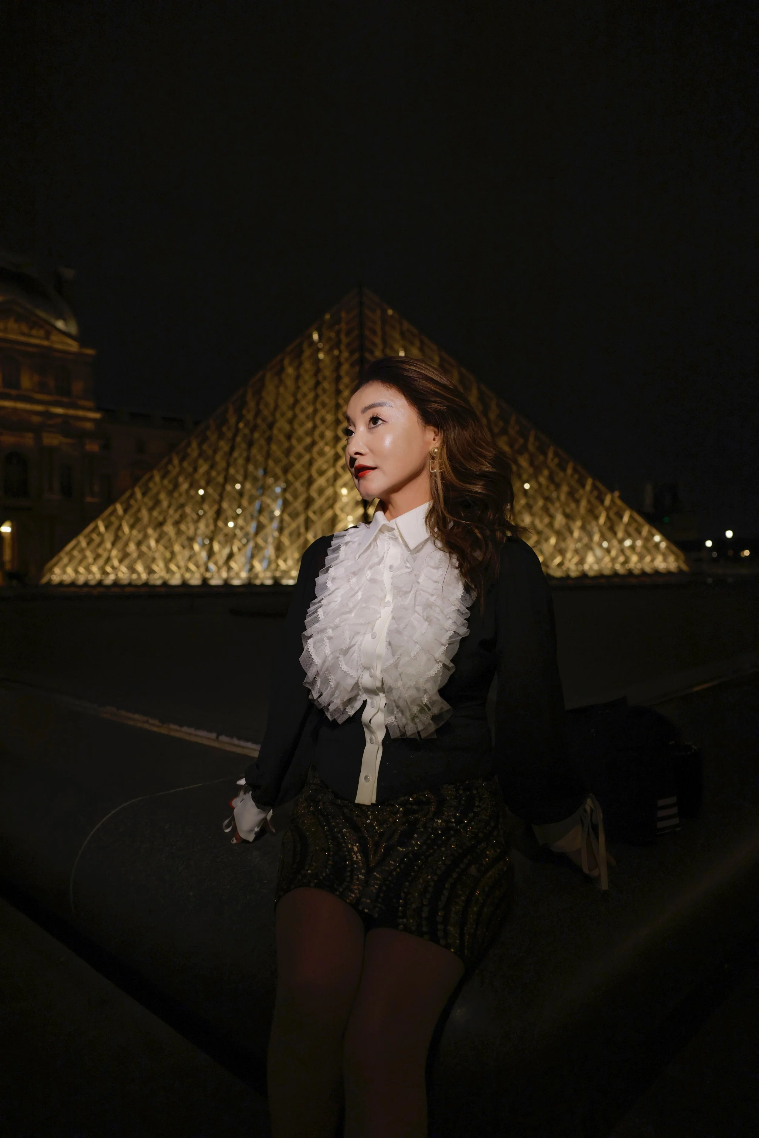 Winnie Wen posing at night in a white ruffled top and trousers standing in front of the Louvre Pyramid, Paris France.