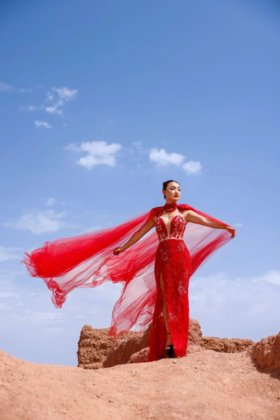 Winnie Wen in a red, elegant, lacy dress with a high slit, standing on a rocky terrain against a blue sky with clouds, with her arms outstretched holding a flowing red cape in the Moroccan desert.