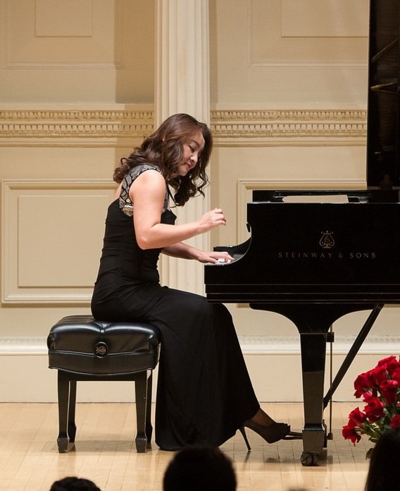 A woman in a black dress playing a Steinway & Sons grand piano on stage, with a beige ornate wall behind her and red flowers in the foreground.