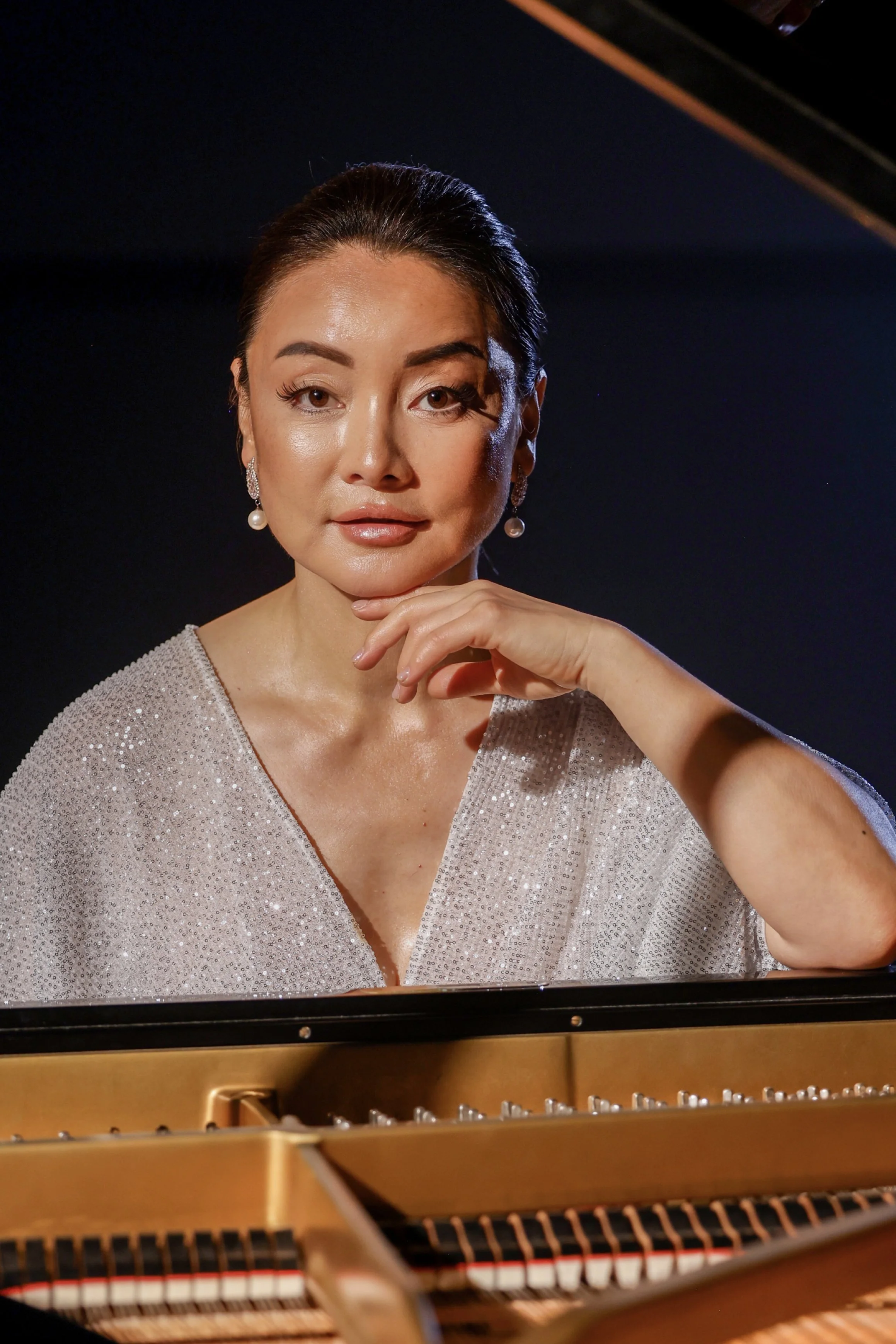Close-up of Winnie Wen wearing pearl earrings and a sparkly, elegant dress, sitting at a grand piano.