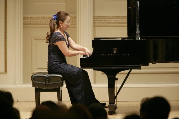 A woman in a dark blue lace dress playing a grand piano on a stage with an audience watching.
