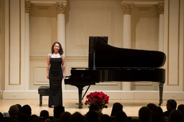 A woman wearing a black dress stands on a stage beside a grand piano, with an audience in front of her and a large bouquet of red flowers in front of the piano.