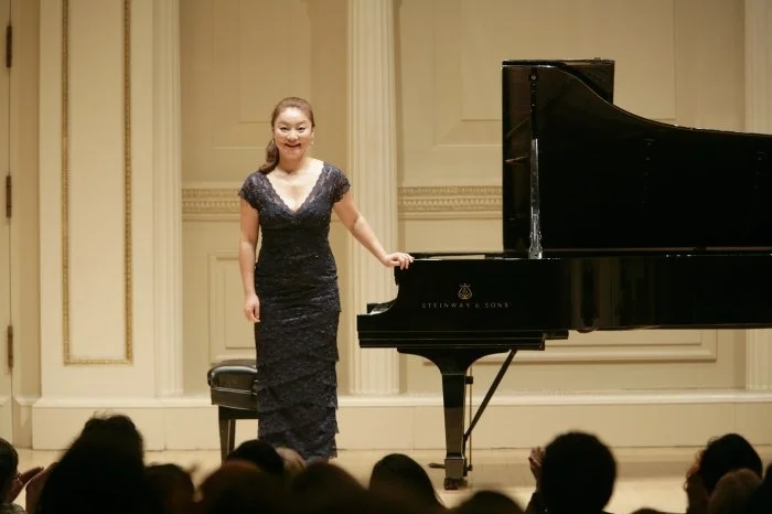 A woman in a black dress standing beside a grand piano on stage, smiling at the audience.