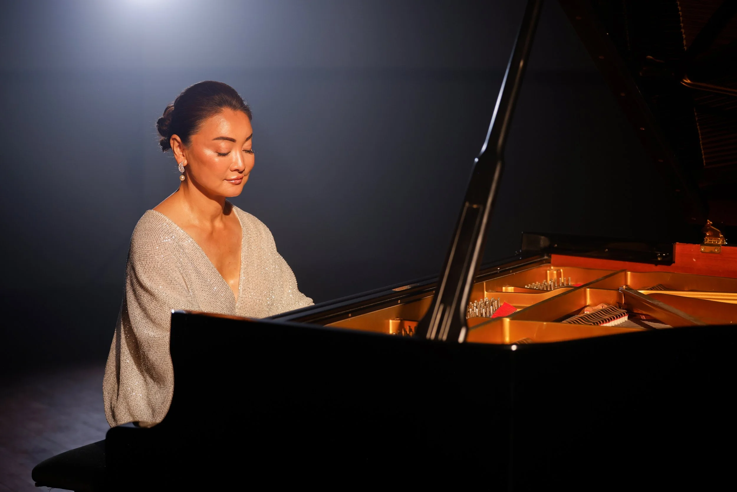Winnie Wen in a shimmering white dress, playing a grand piano against a dark background with a spotlight on her.