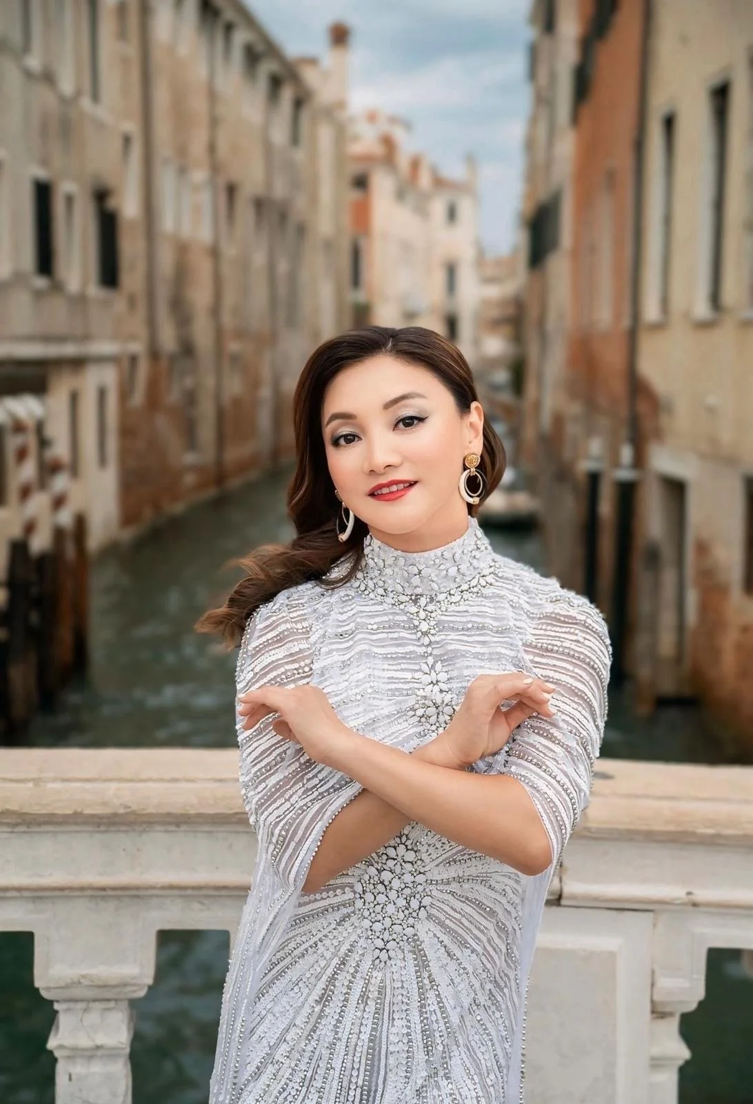 Winnie Wen wearing a white, embellished dress, standing on a bridge in Venice with a canal and historic buildings in the background.
