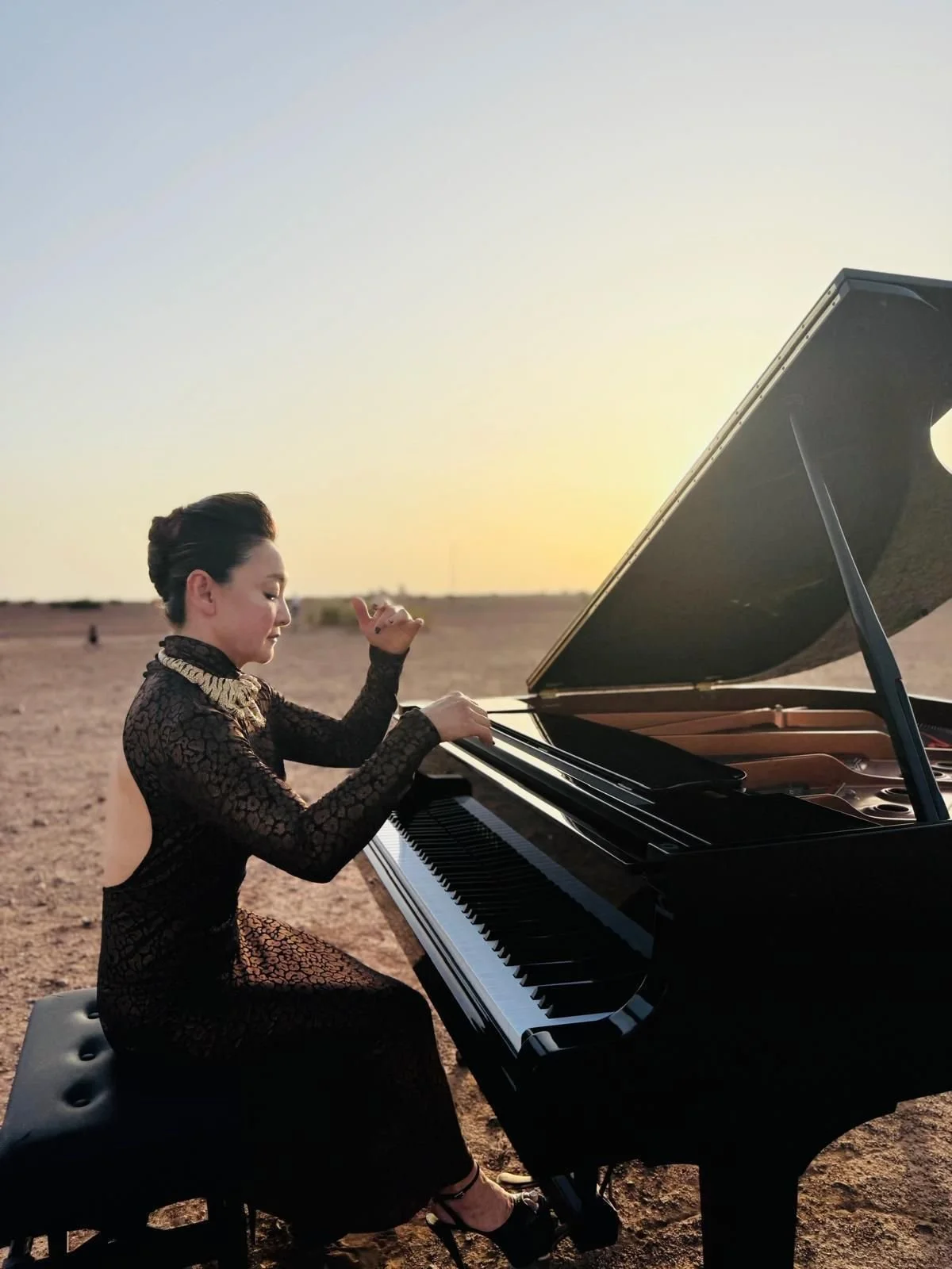 Winnie Wen in a black lace dress with a high collar, sitting at a grand piano outdoors during sunset in the Moroccan desert.