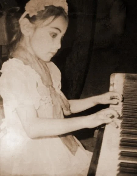 A young girl with light-colored hair and styled with flowers in her hair is playing a piano, dressed in a light-colored dress with lace details.