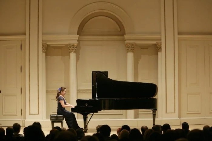 Young girl playing the piano on stage at a concert hall with an audience watching.