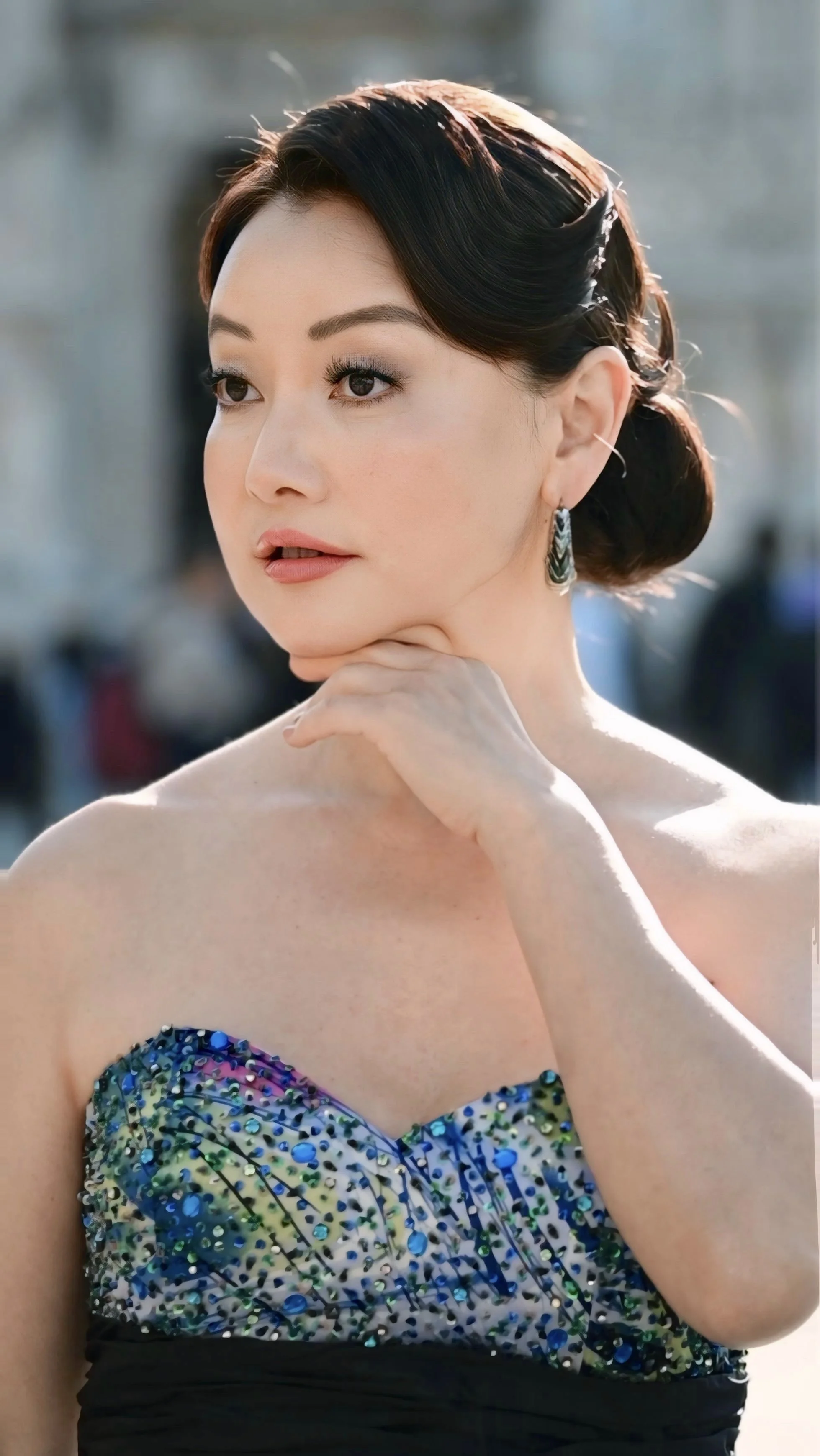 Close-up of Winnie Wen styled in an elegant updo, wearing makeup, earrings, and a strapless dress with a colorful, sequined pattern on the bodice, outdoors with the Duomo di Milano in the background.