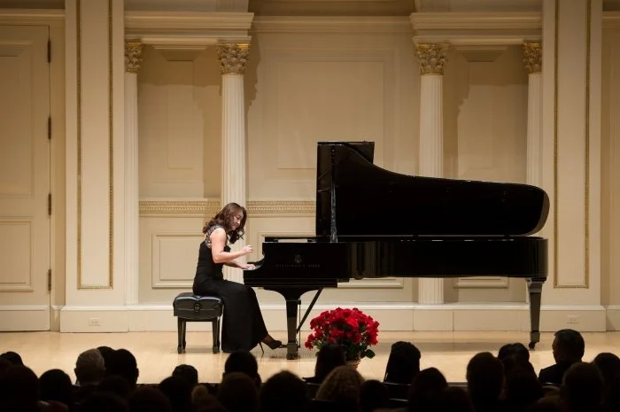 Winnie Wen on stage in a black dress playing a grand piano at Carnegie Hall with an audience watching.