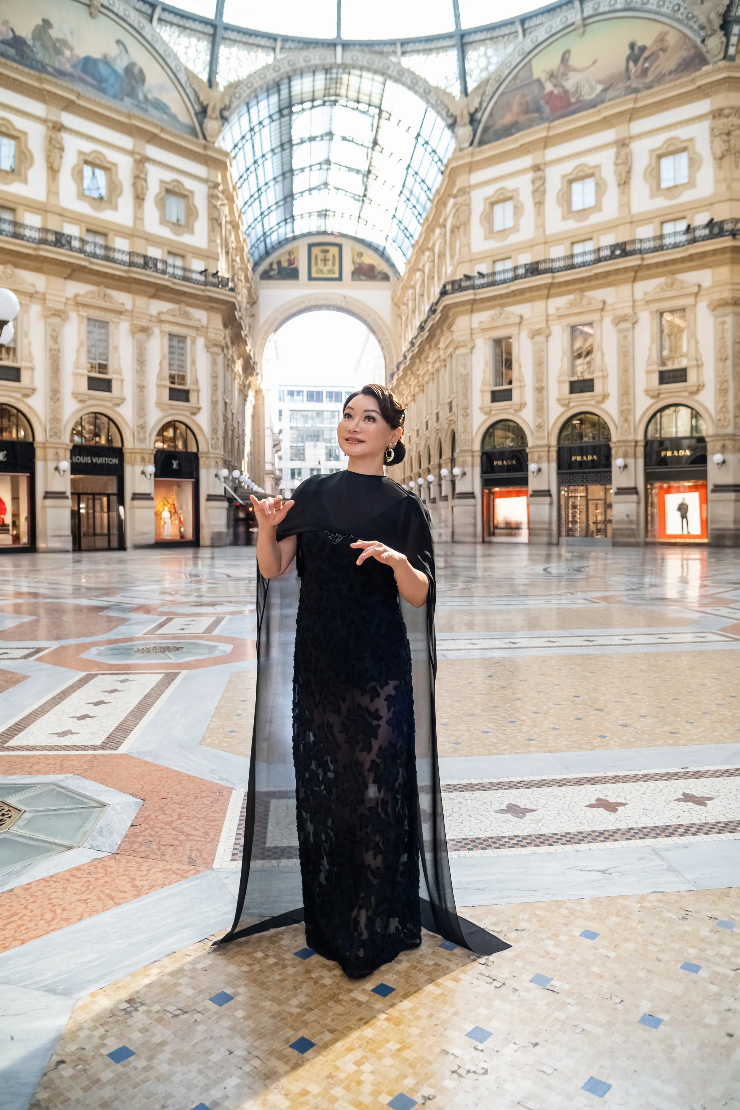 Winnie Wen in a black dress and chandelier earrings stands inside Galleria Vittorio Emanuele II with high glass ceilings, ornate architecture, and luxury store displays in the background.