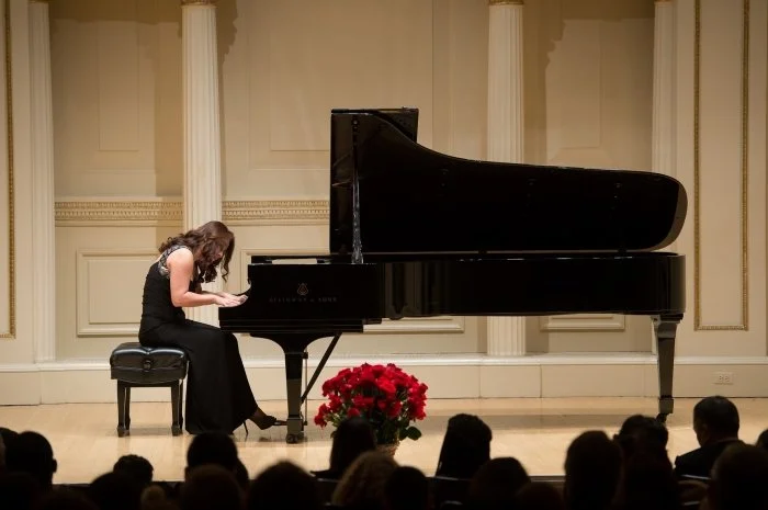 A woman playing a grand piano on a stage during a performance or recital, with an audience in front.