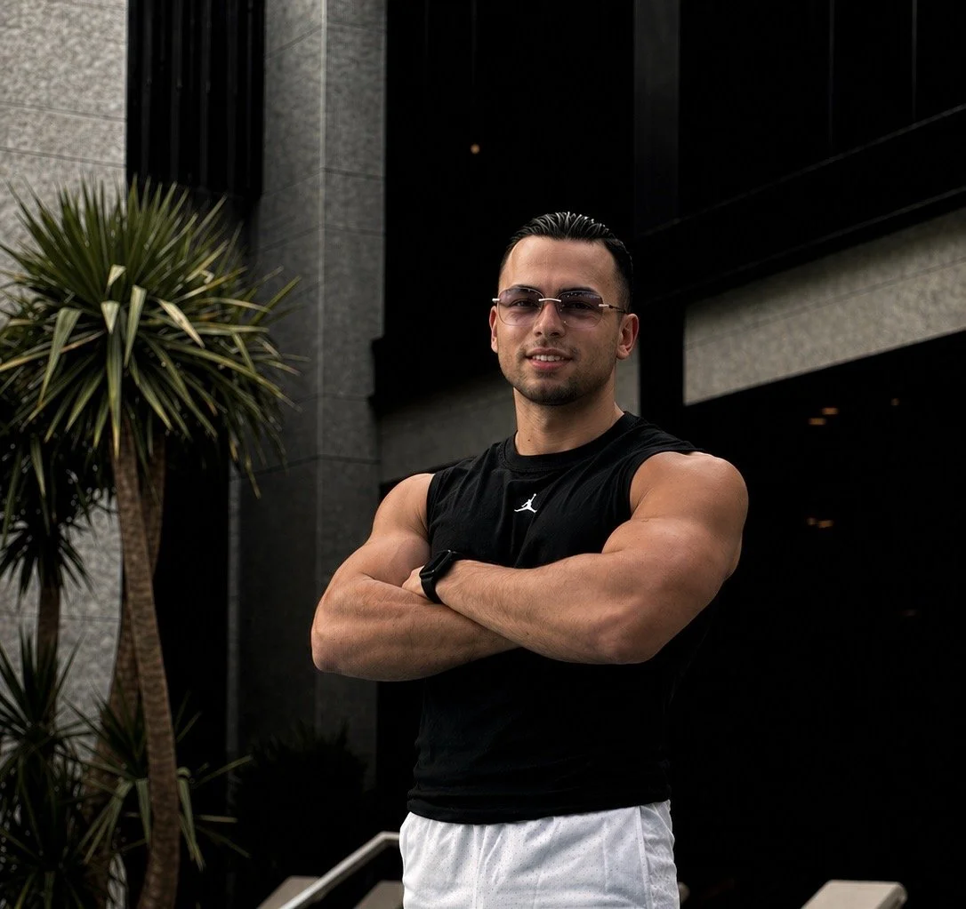 Junger Mann mit Sonnenbrille, trainiert, steht vor dunklem Gebäude, trägt schwarze Basketballweste, weiße Shorts, in der Sonne, mit Armschonern.