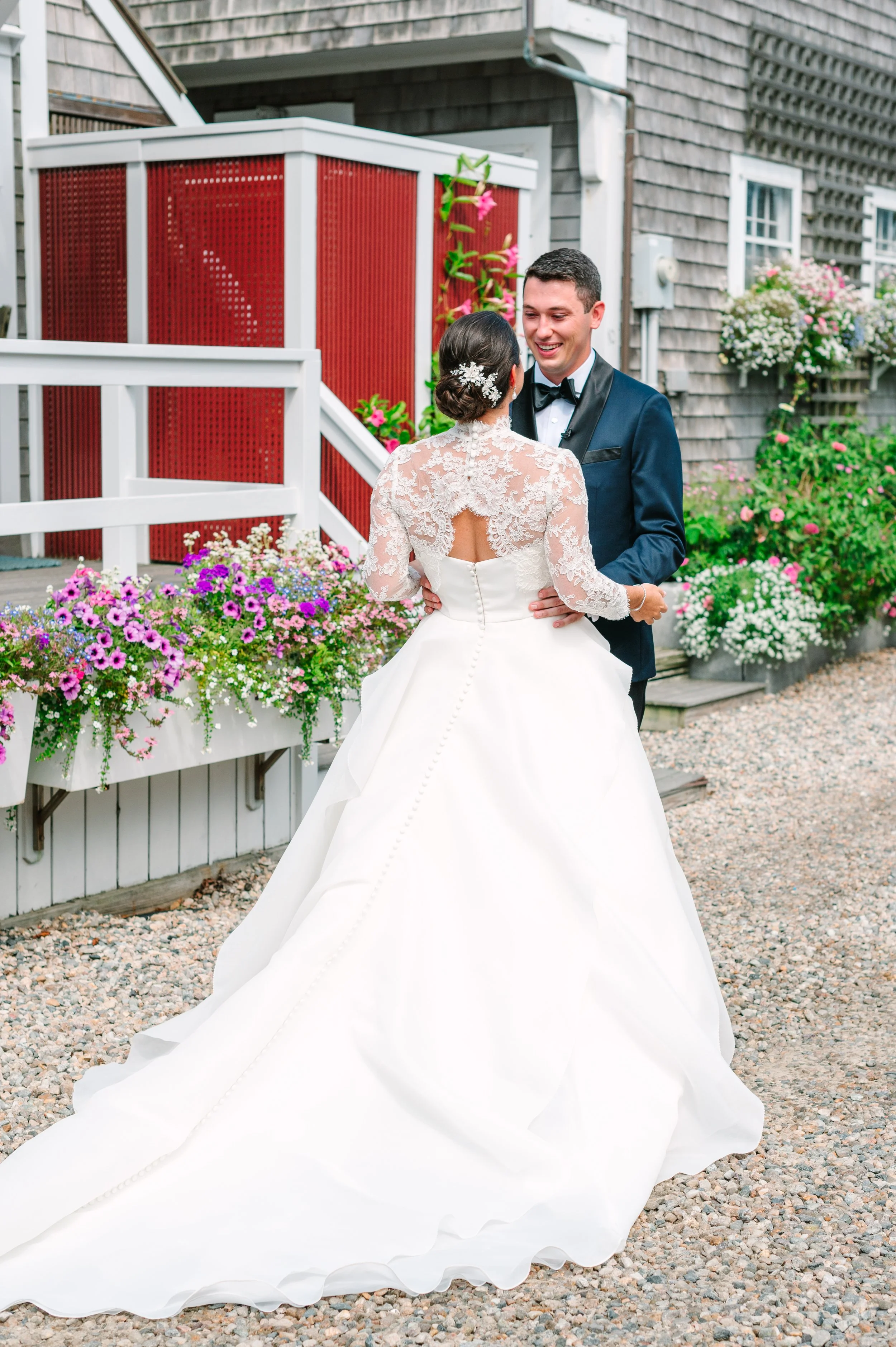 Nantucket wedding first look. A bride and groom smile at each other outdoors, surrounded by colorful flowers and wooden buildings, during a wedding celebration.