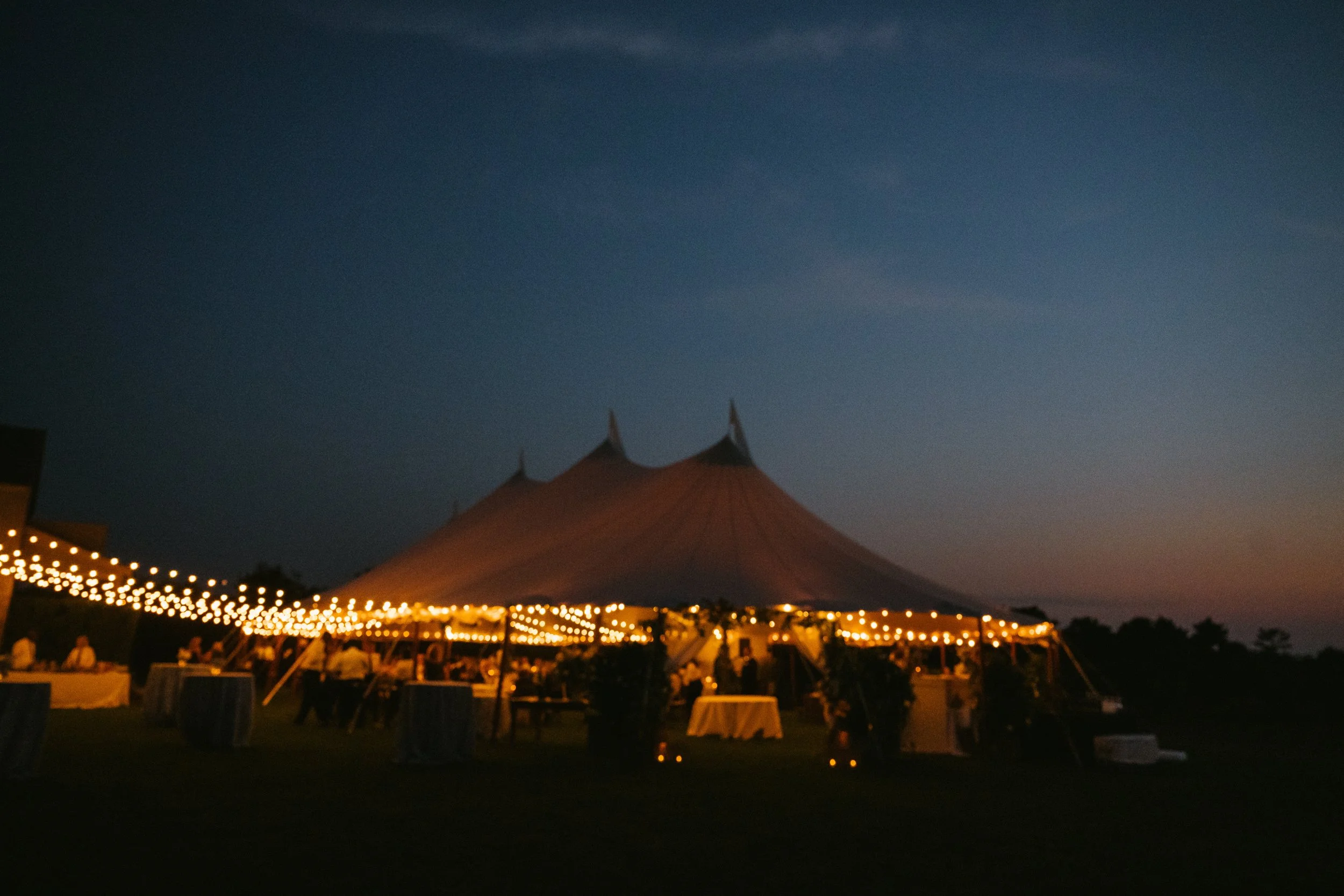 Nantucket tented wedding. Night scene of outdoor event with string lights, tables, and a large tent in the background.