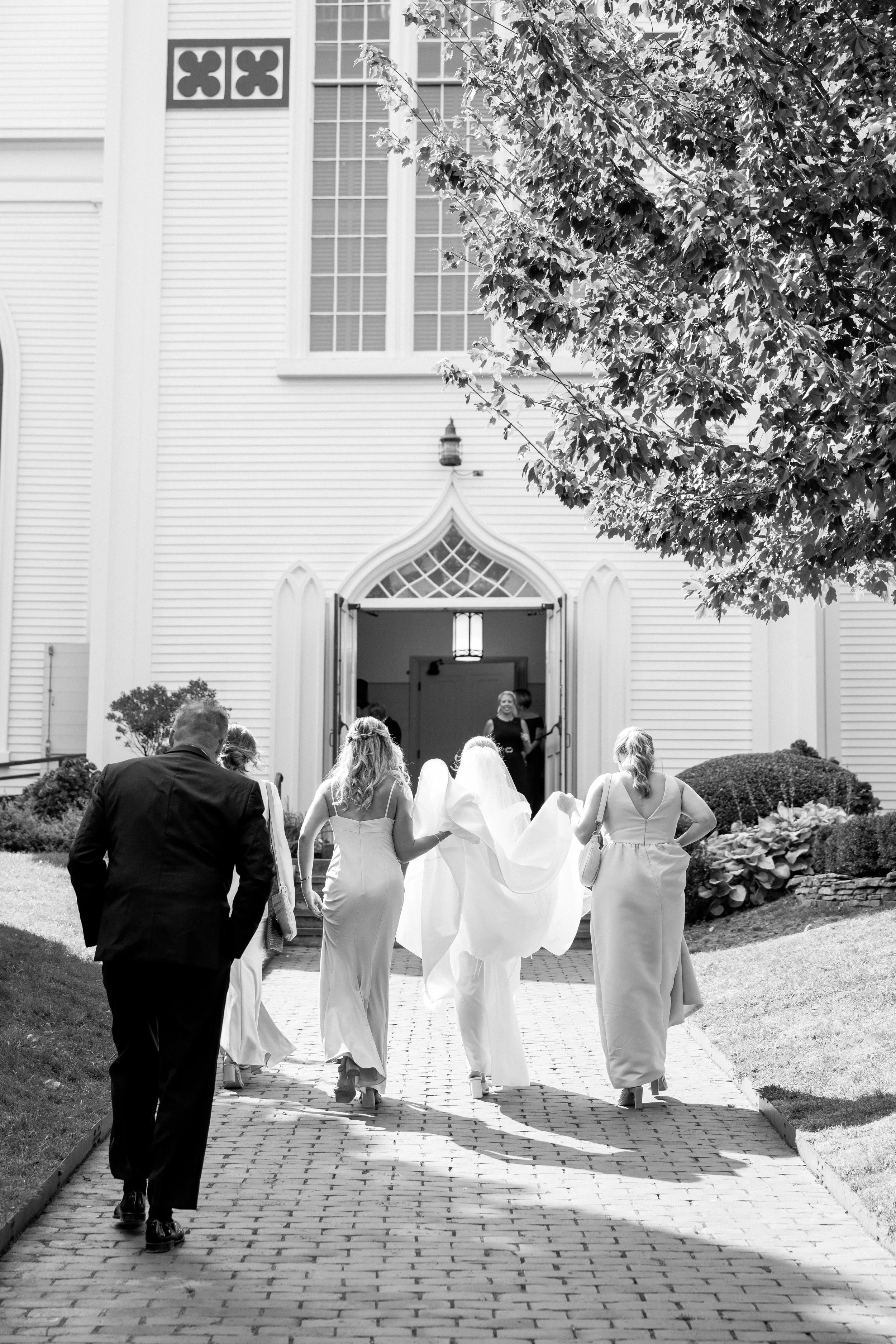 Nantucket wedding ceremony. Black and white photo of a wedding party walking out of a church, with bridesmaids in dresses and a bride in a gown, followed by a man in a suit, on a brick path with a large tree and shrubs in the background.
