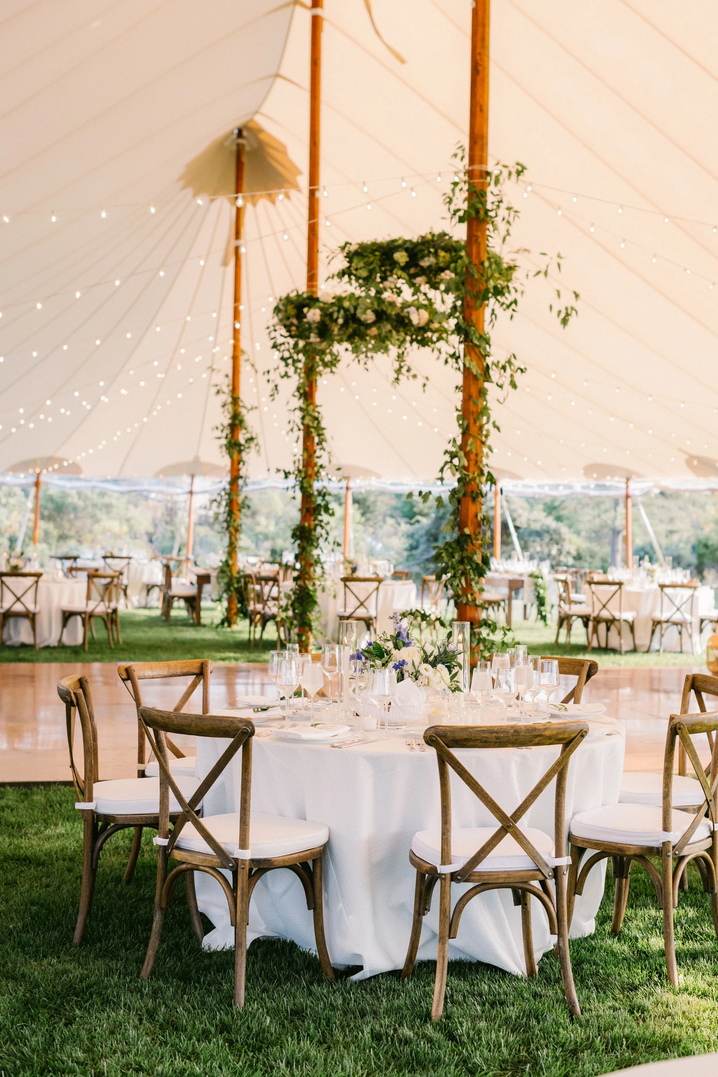 Nantucket tented wedding. Elegant outdoor wedding reception setup with round tables, white tablecloths, floral centerpieces, wooden chairs, and a tent decorated with string lights and greenery.