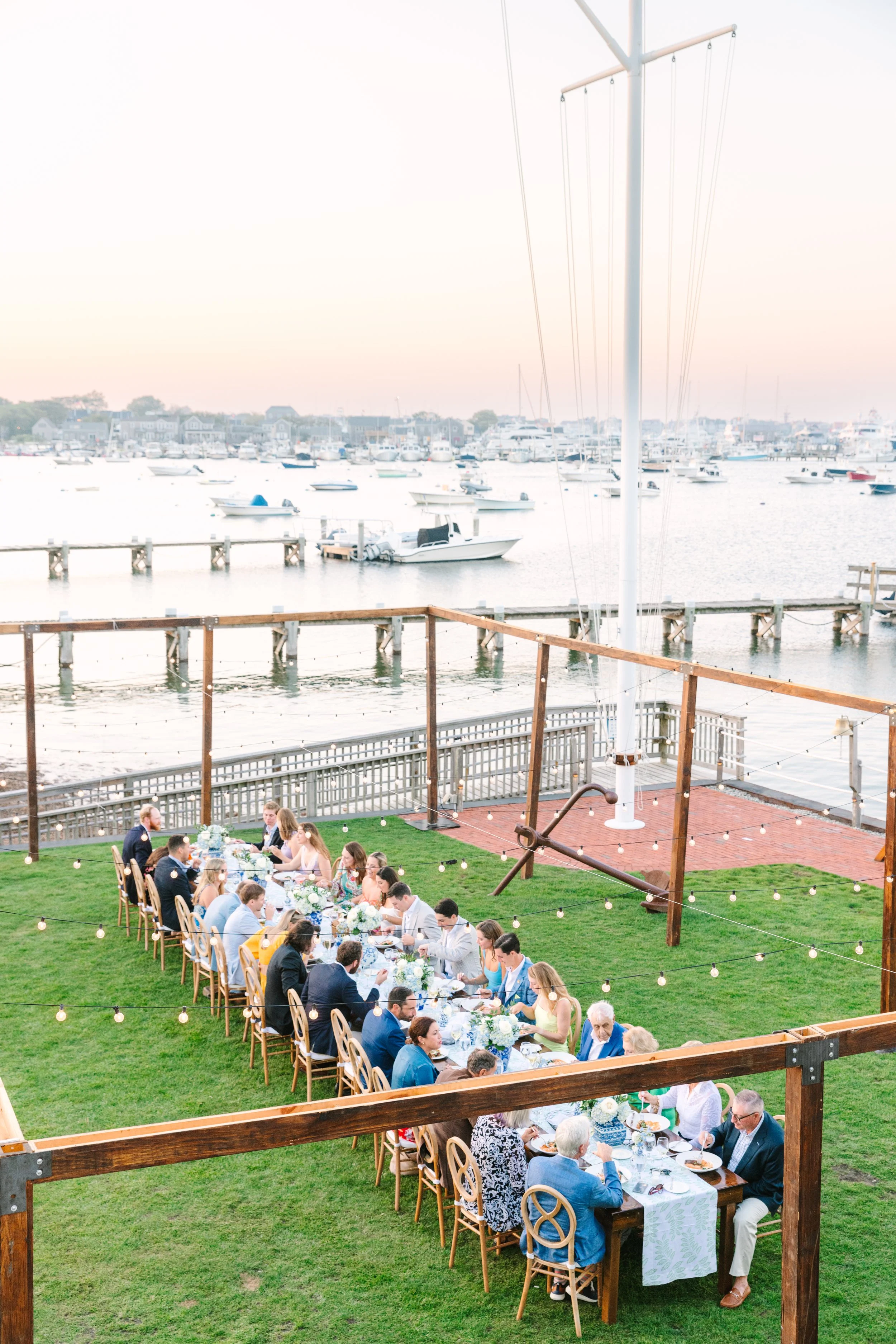 Great Harbor Yacht Club rehearsal dinner.A large outdoor meal gathering on a grassy area near a waterfront with docked boats and a marina in the background. The long table is decorated with flowers and surrounded by people dining under string lights.