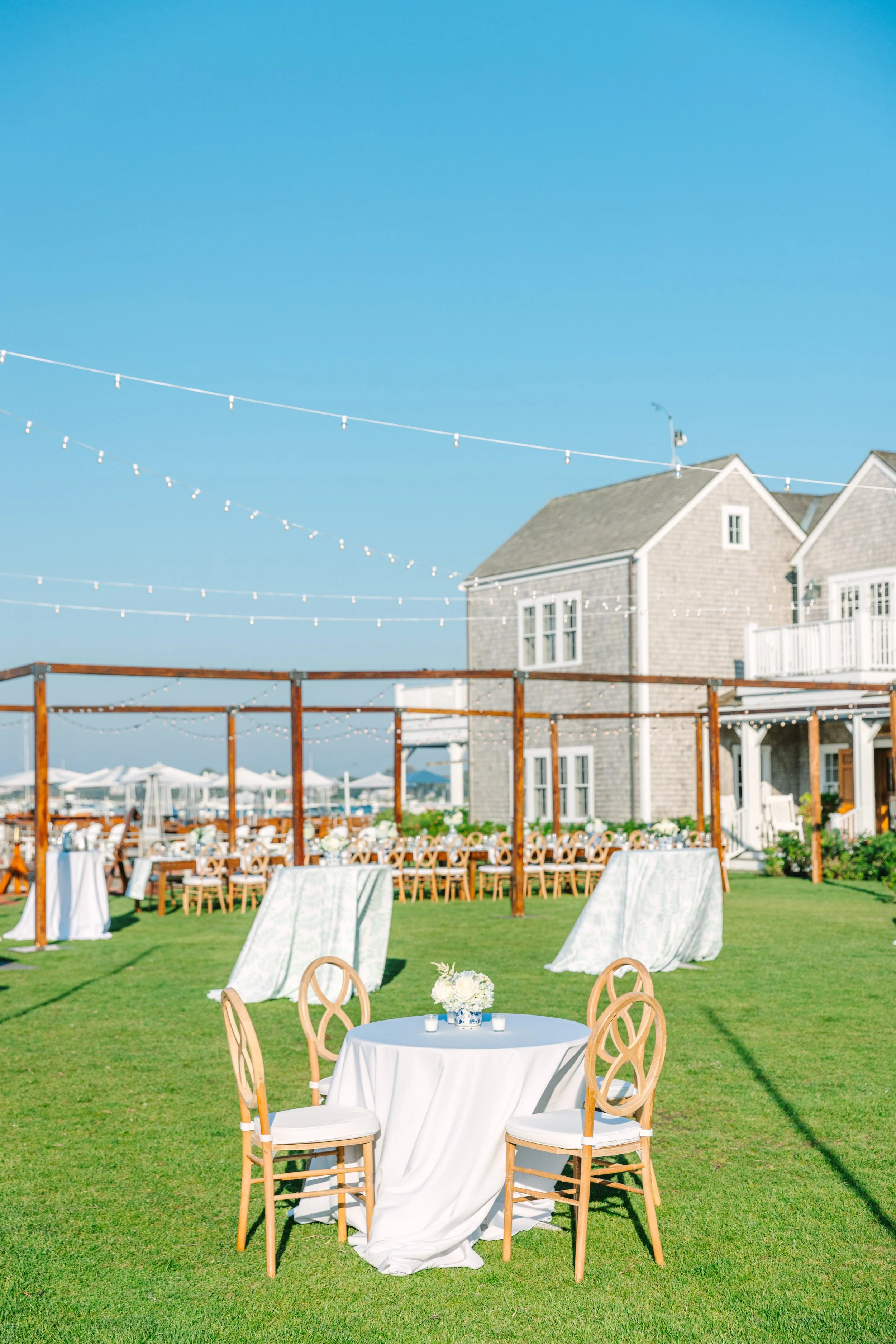 Great Harbor Yacht Club Welcome Party. Outdoor wedding reception setup on a grassy lawn with tables and chairs, decorated with white tablecloths and flowers, under string lights, with a large house and beach umbrellas in the background.