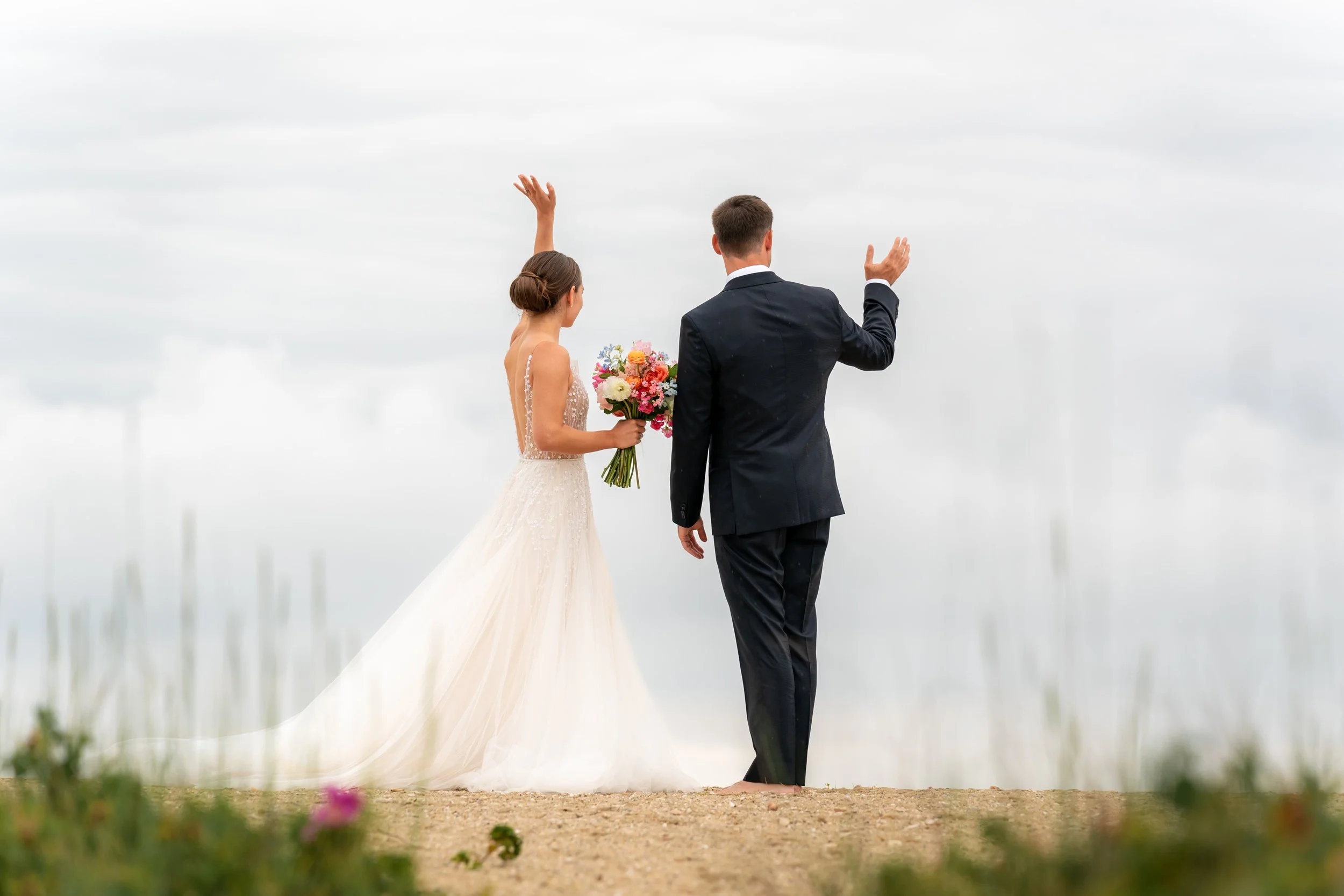 Brant Point Nantucket Wedding. A bride and groom standing outdoors on a sandy surface, facing away from the camera, with the bride holding a bouquet of flowers and the groom raising his hand, during a wedding ceremony.